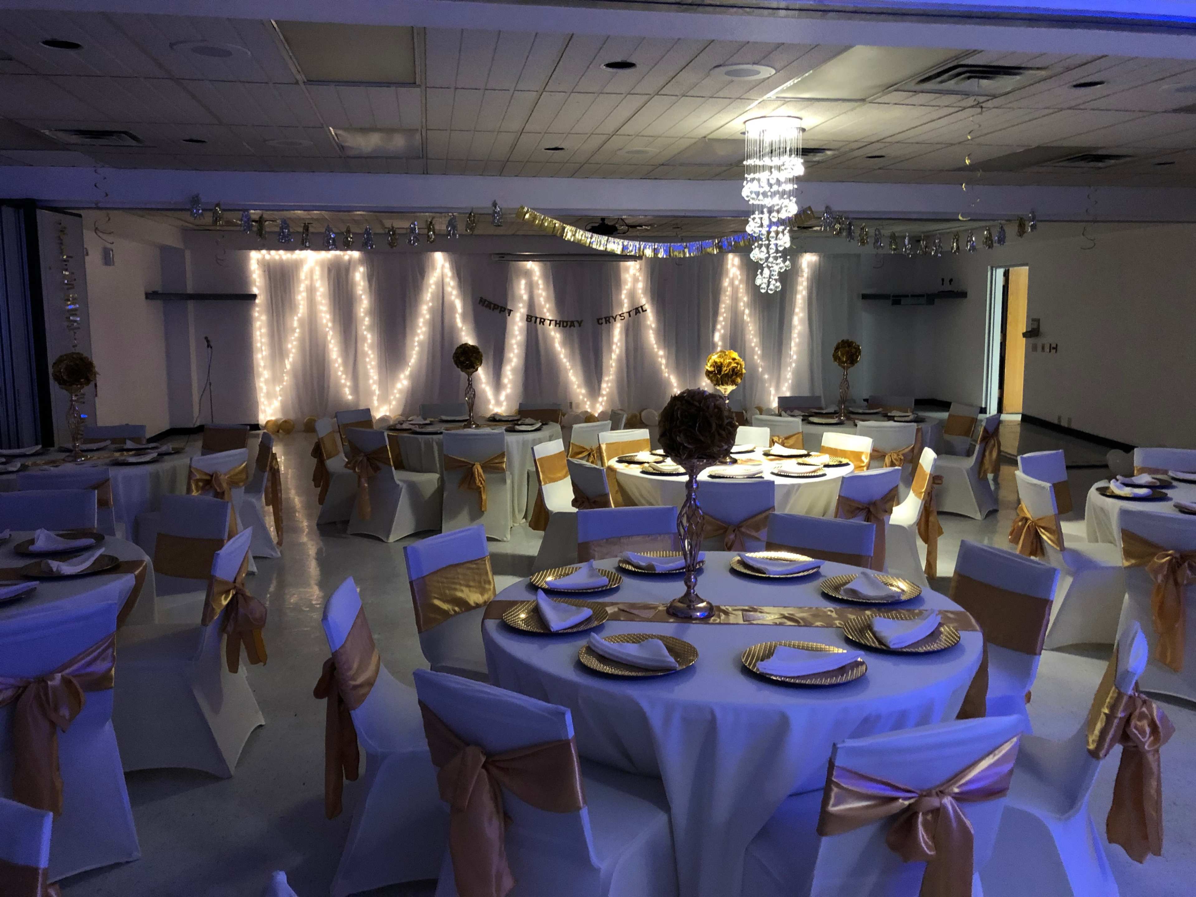 A decorated hall set up for an event, featuring round tables with white tablecloths and gold accents, surrounded by string lights and ornamental centerpieces.