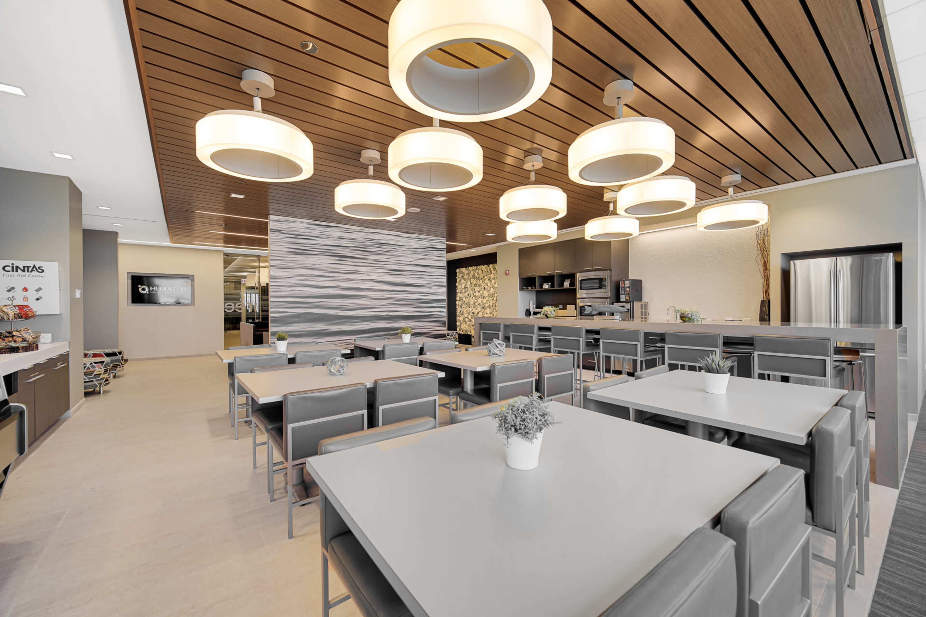 The image shows a modern dining area with several tables and chairs, illuminated by large pendant lights, and a kitchen in the background.