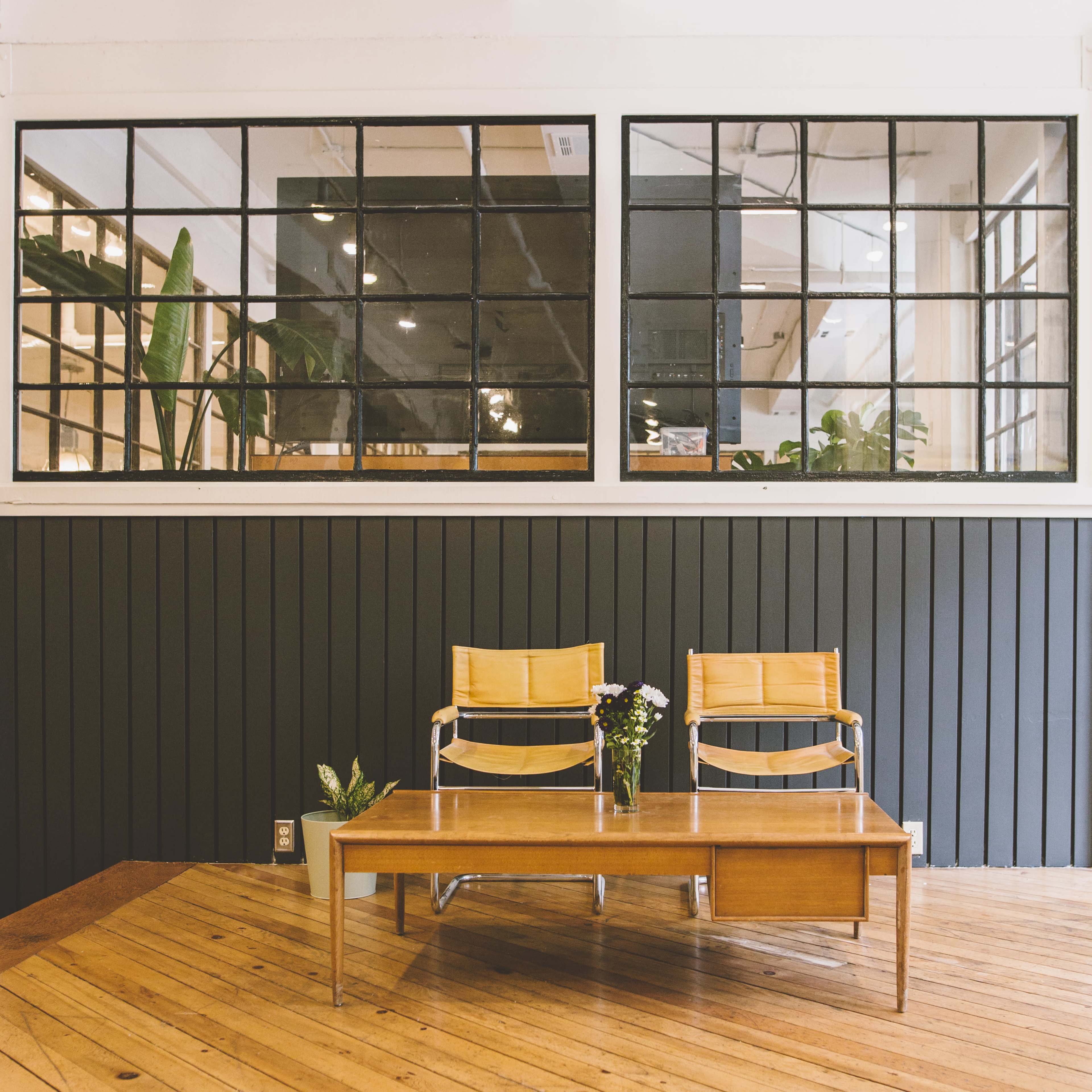 A minimalist interior with two yellow chairs next to a wooden table, set against a dark wall and large windows featuring plants.