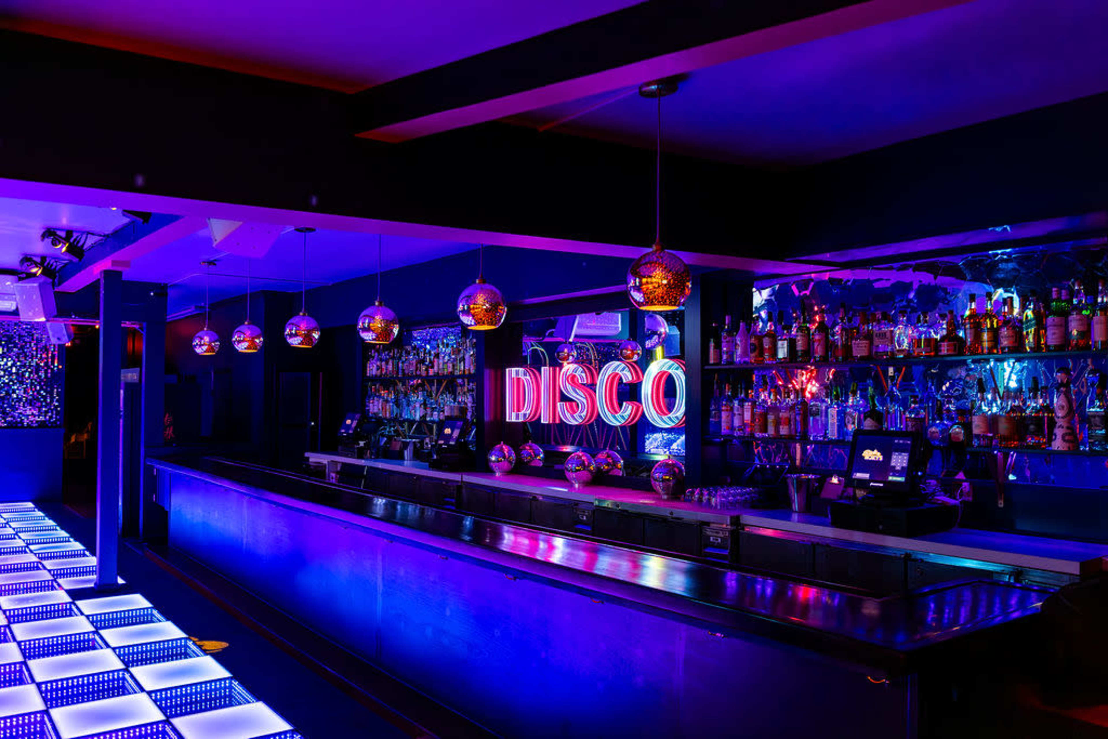 A modern bar features a brightly lit "DISCO" sign, with colorful LED lights illuminating the space and a long countertop lined with liquor bottles.