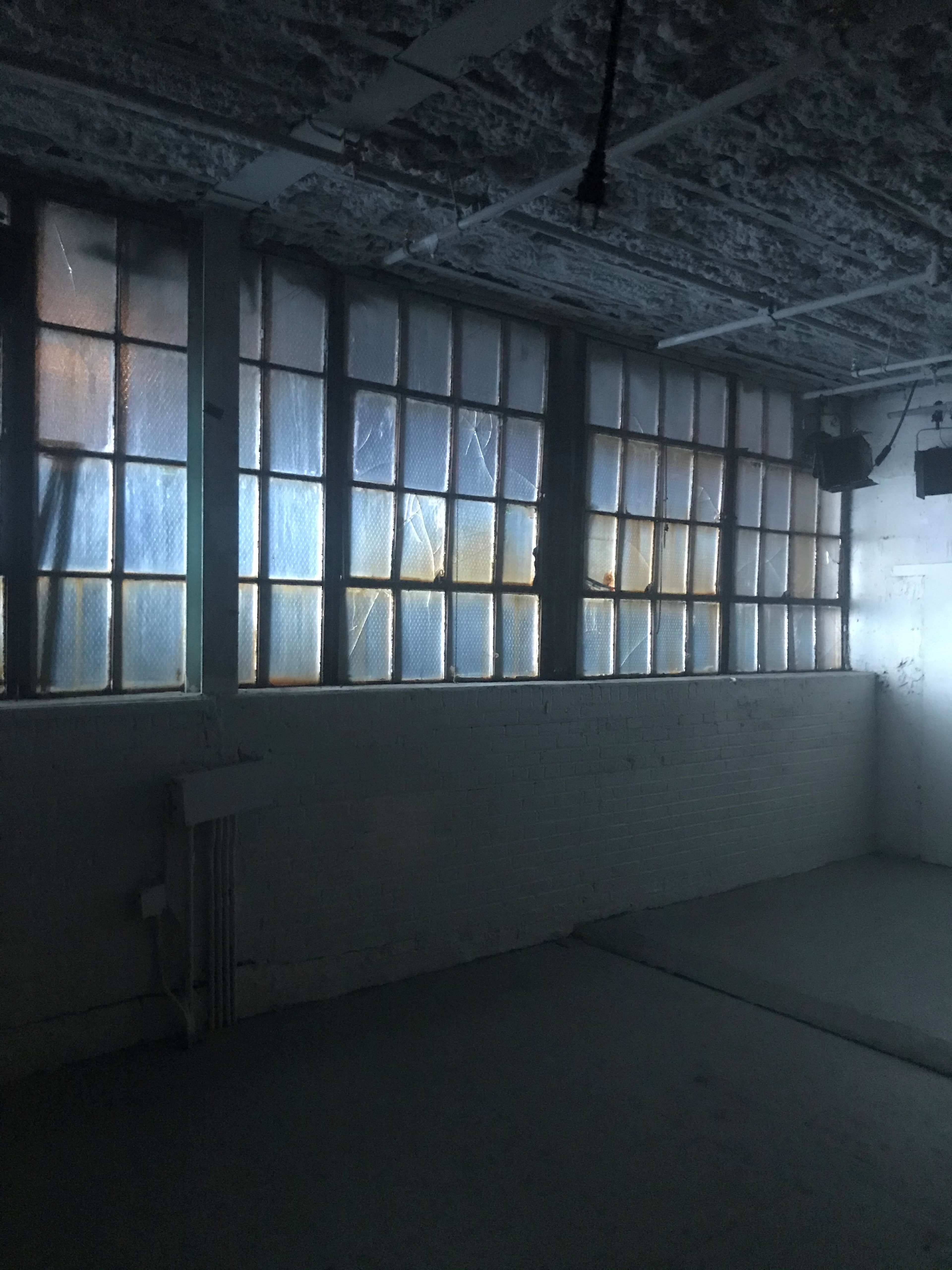A dimly lit interior space with large, multi-paned windows covered by blue material, casting soft light onto a bare floor.