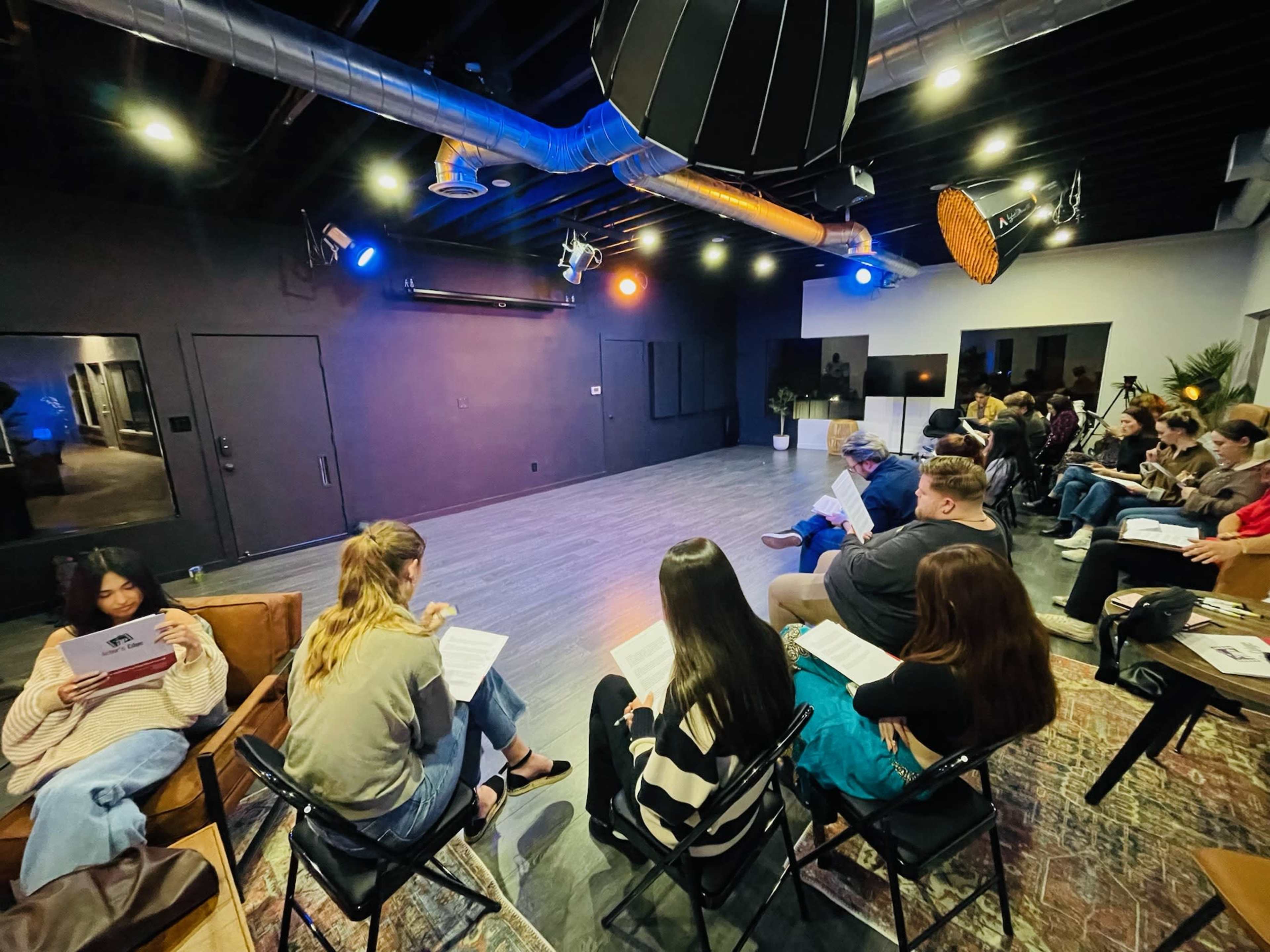 A group of people sits in a rehearsal space, facing a stage with soft lighting, while some attendees hold scripts and notepads.