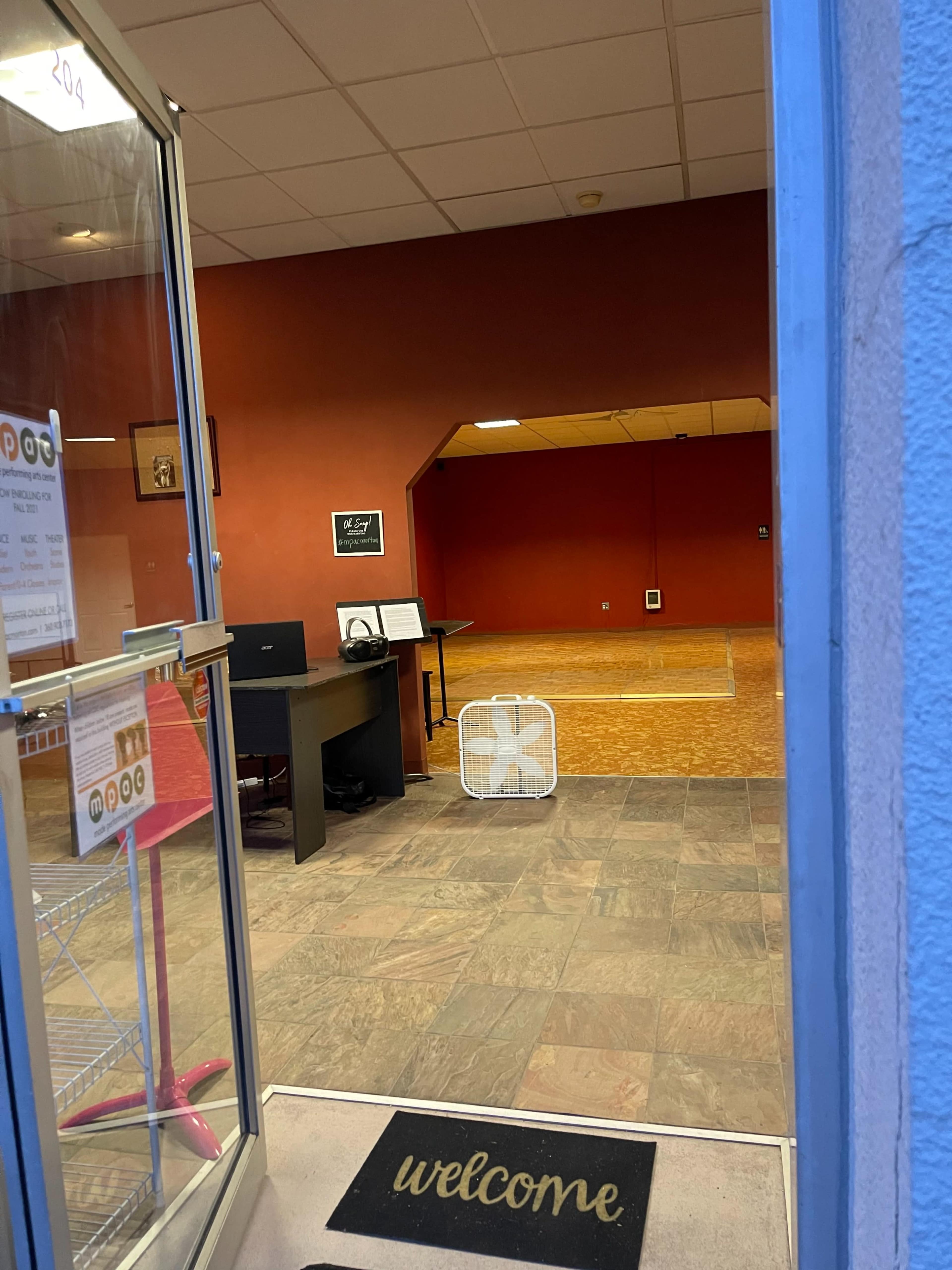 An entrance through a glass door leading into a room with a desk, a fan on the floor, and an empty space with a red wall in the background.