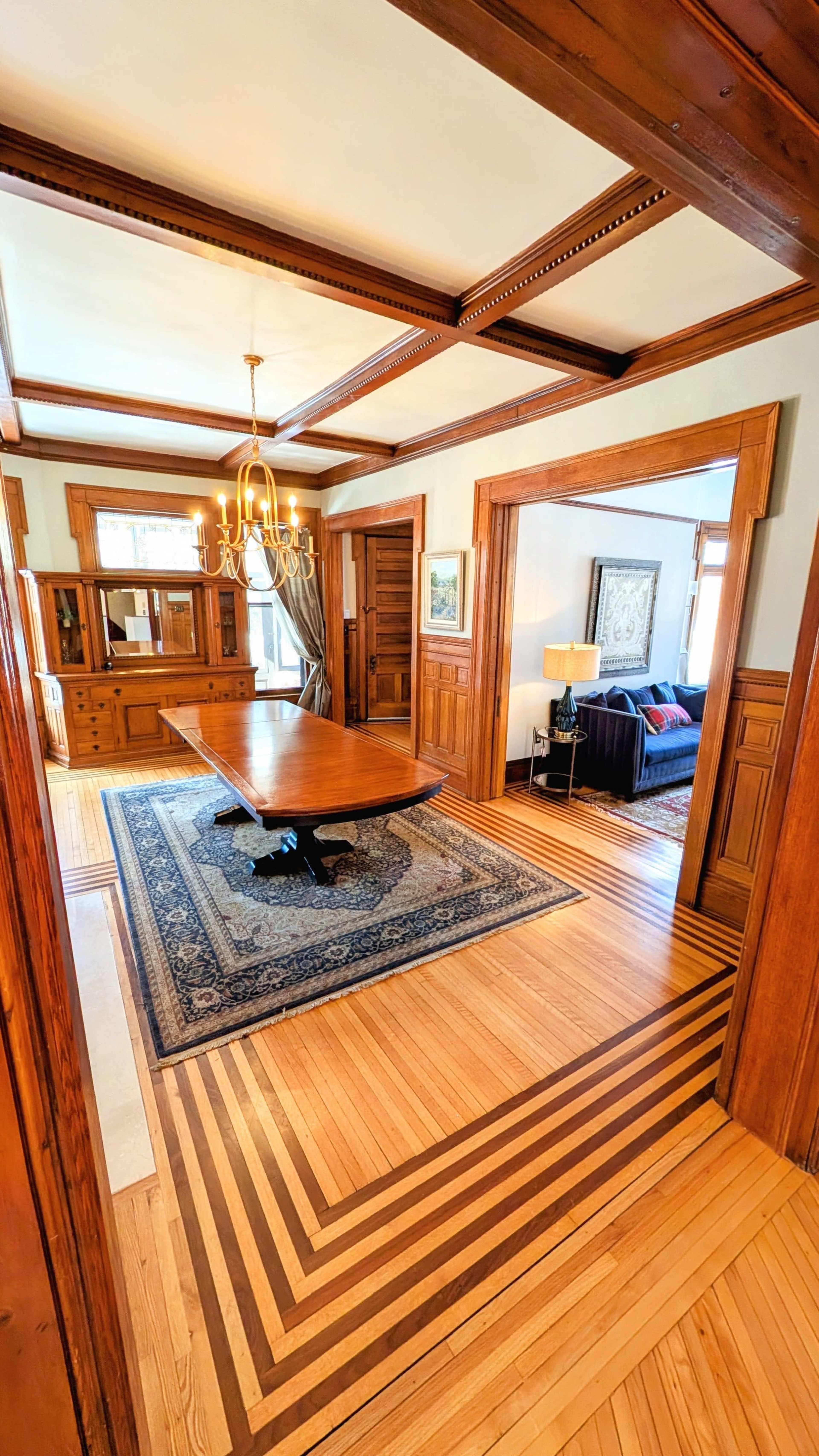 The image shows a spacious dining room featuring a large wooden table, a chandelier, and intricately patterned wooden flooring.