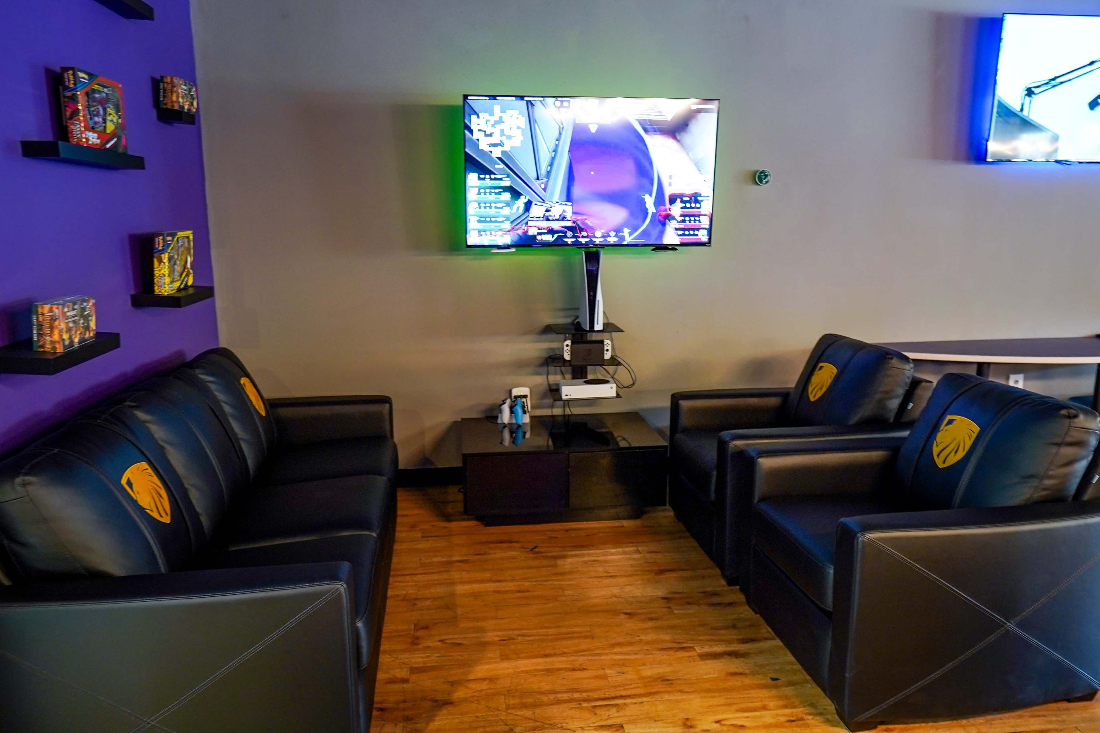A seating area features two black leather chairs with golden emblems, a television mounted on the wall displaying a gaming stream, and a coffee table in between.