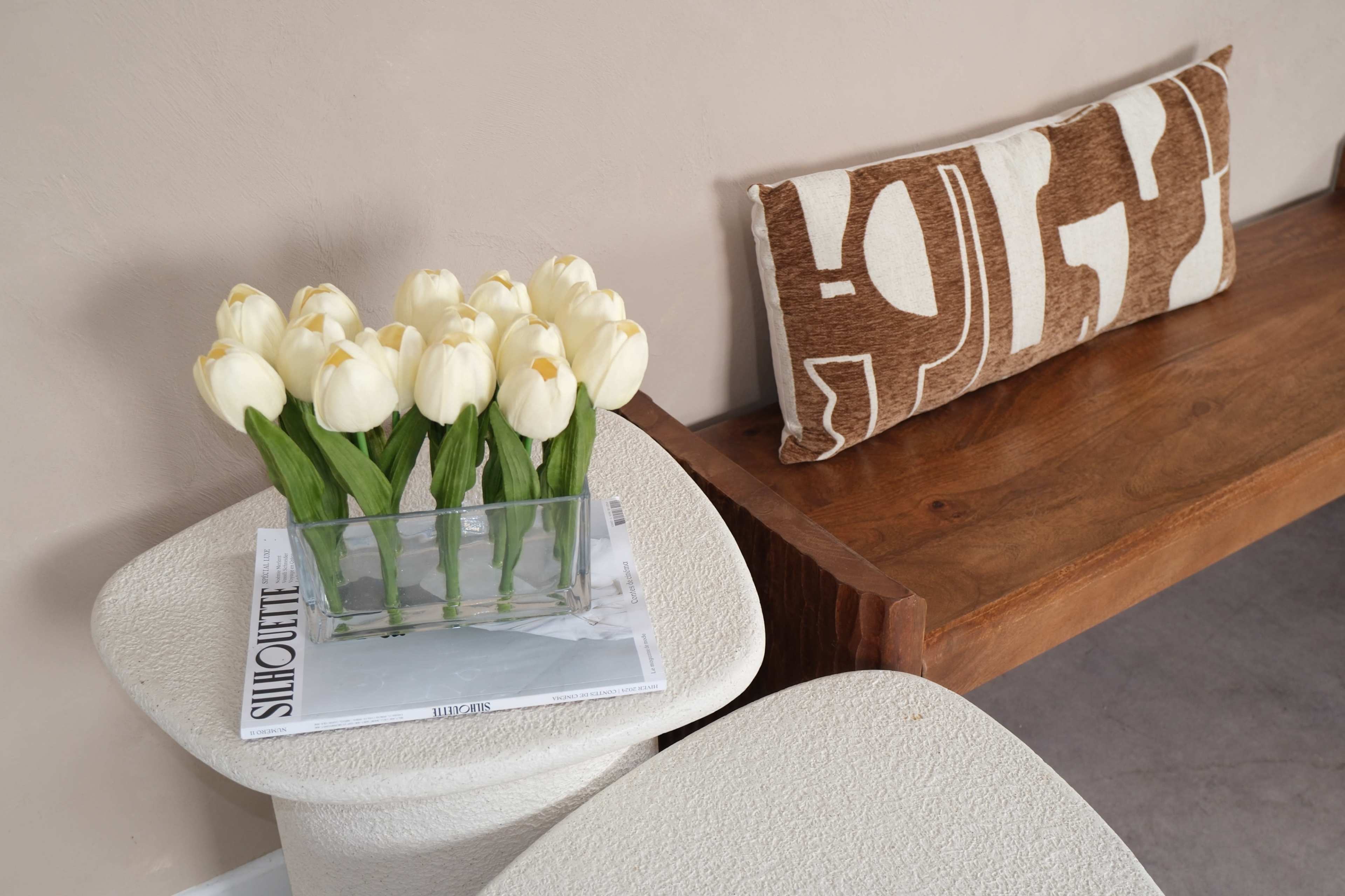 A vase of white tulips sits on top of a magazine placed on a textured side table next to a wooden bench with a patterned pillow.