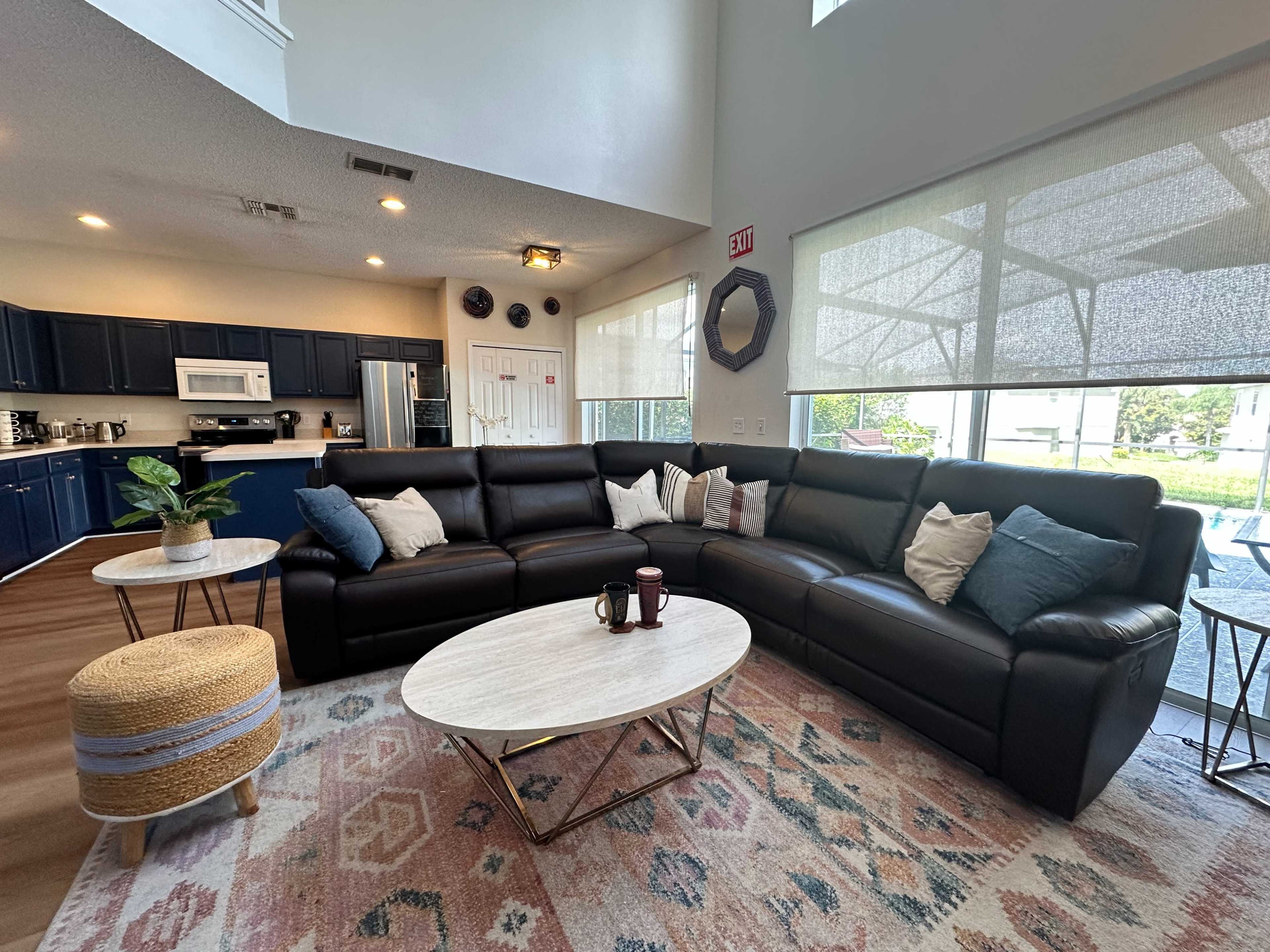 The image shows a modern living room with a large black sectional sofa, a round coffee table, and a patterned rug, all arranged around a bright window that overlooks an outdoor area.