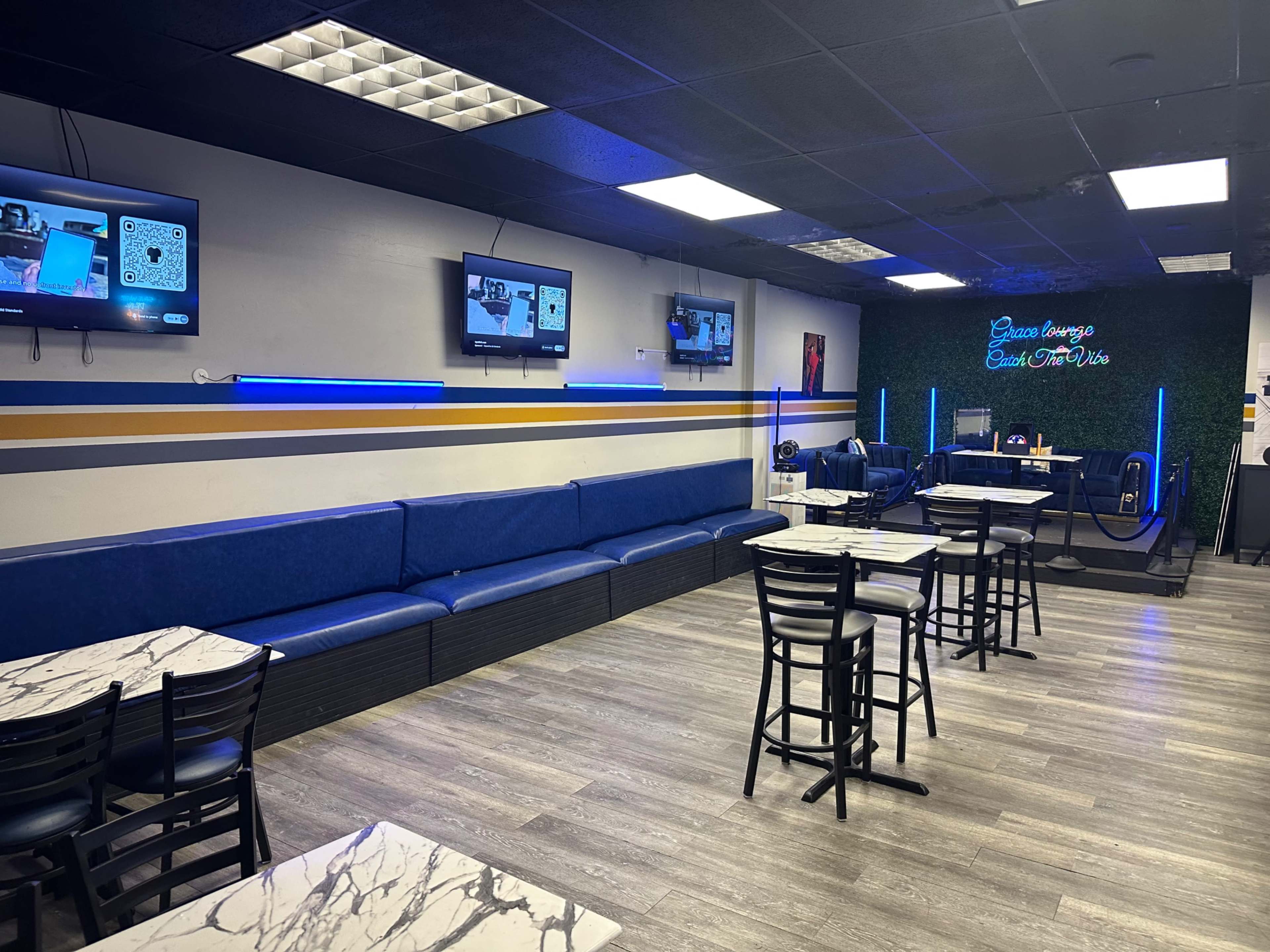 The image shows a modern cafe with blue seating, marble-topped tables, and several televisions mounted on the walls.