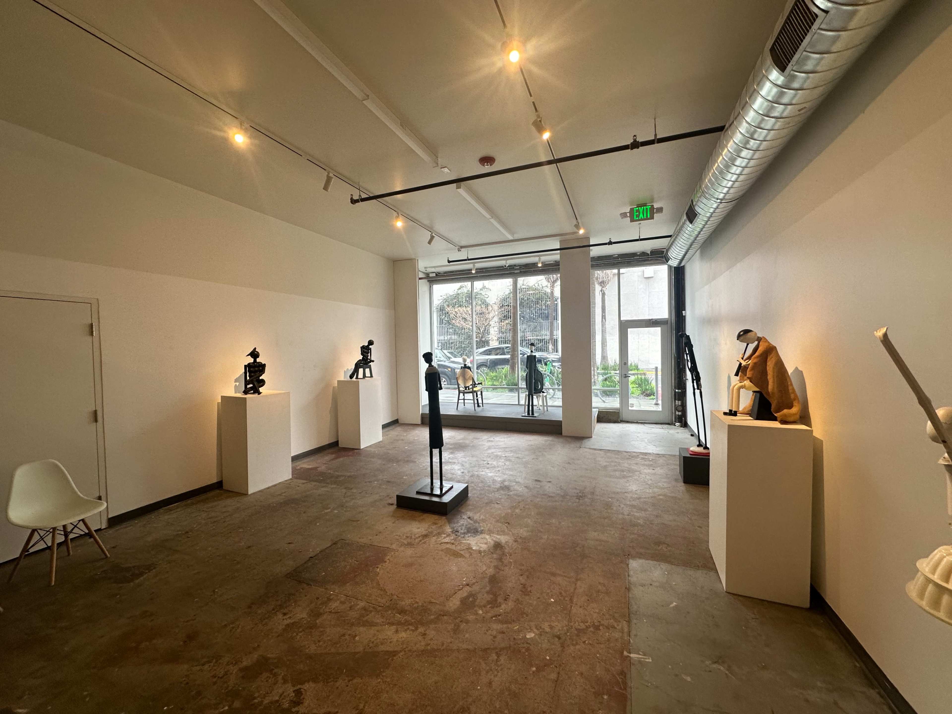 A spacious art gallery featuring several sculptures displayed on pedestals against white walls and a view of an outdoor area through large windows.