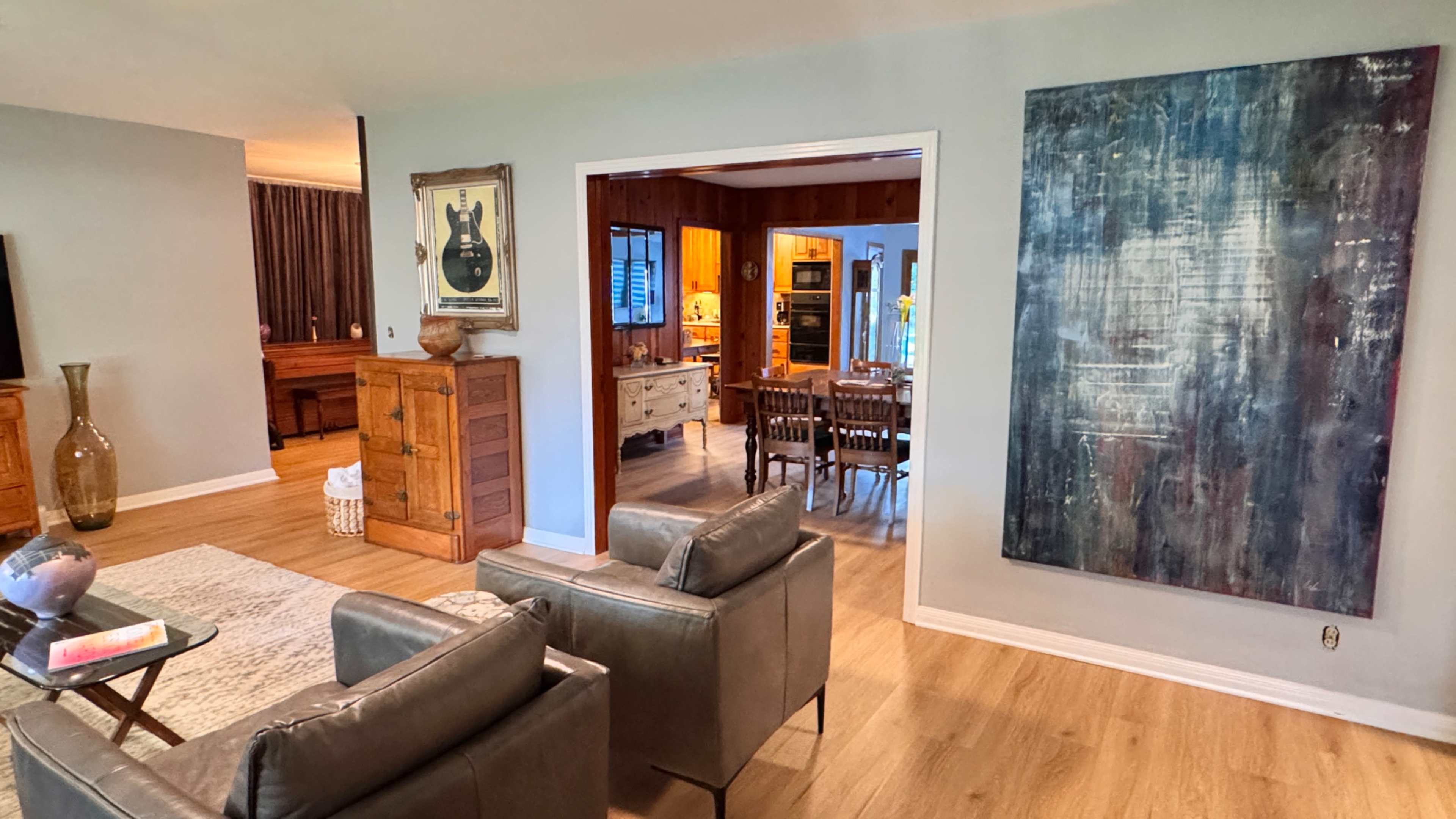 The image shows a spacious living area with modern furniture, a large abstract painting on the wall, and a view into a dining area.