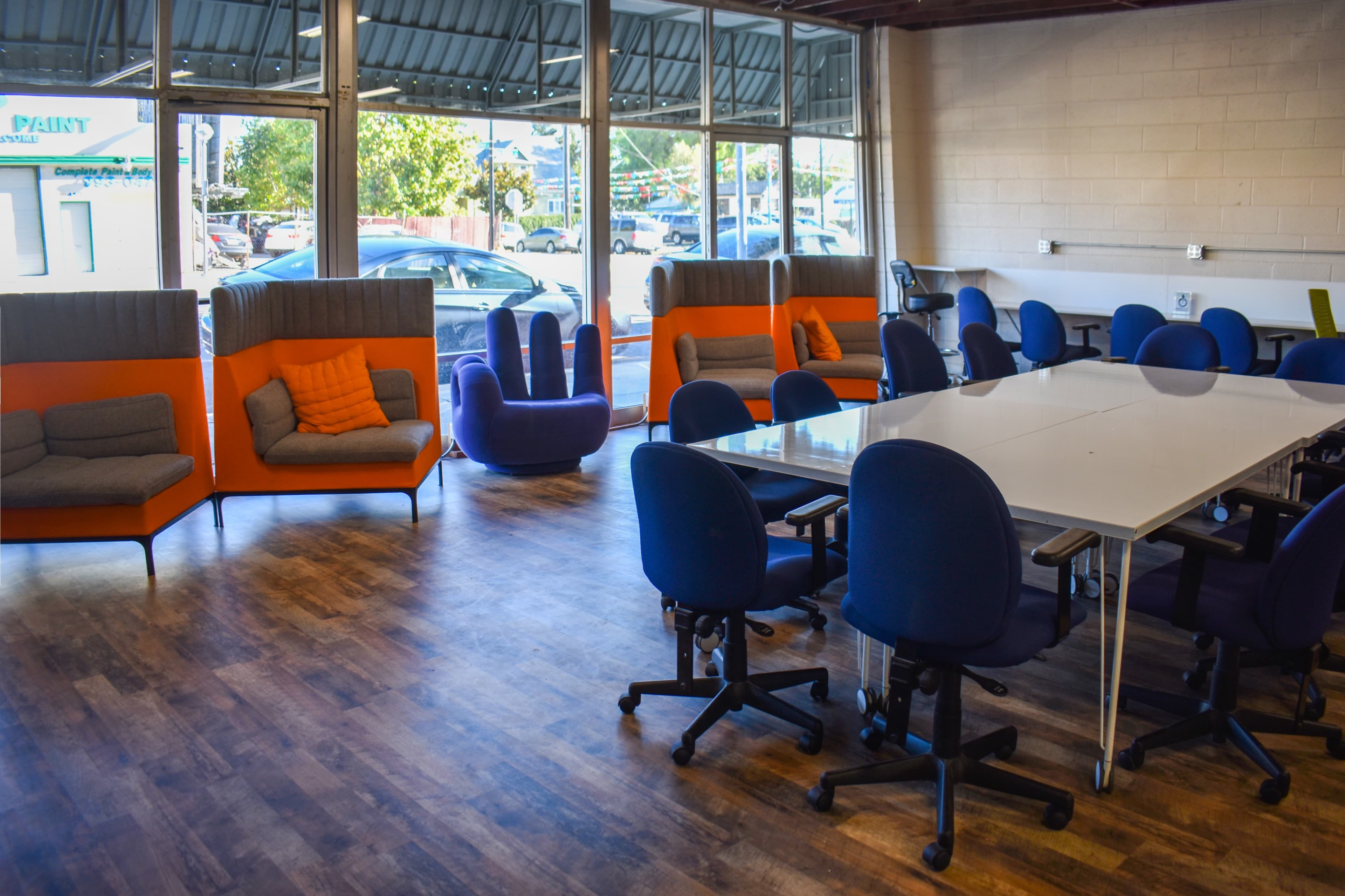The image shows a spacious modern office with a long white table surrounded by blue chairs, and orange-accented seating areas along the windows.