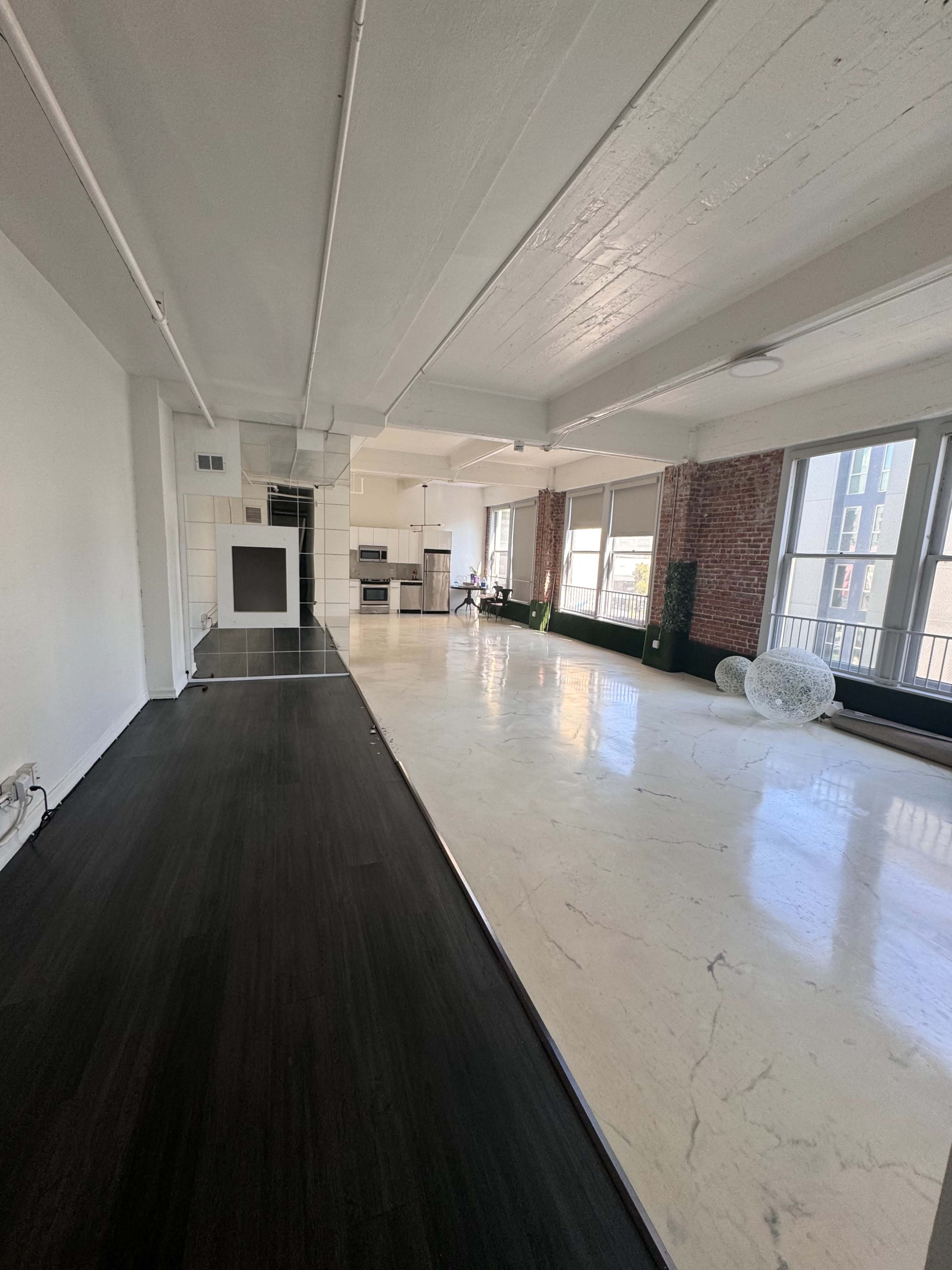 A spacious, modern apartment with a combination of dark and light flooring, large windows, and exposed brick walls.