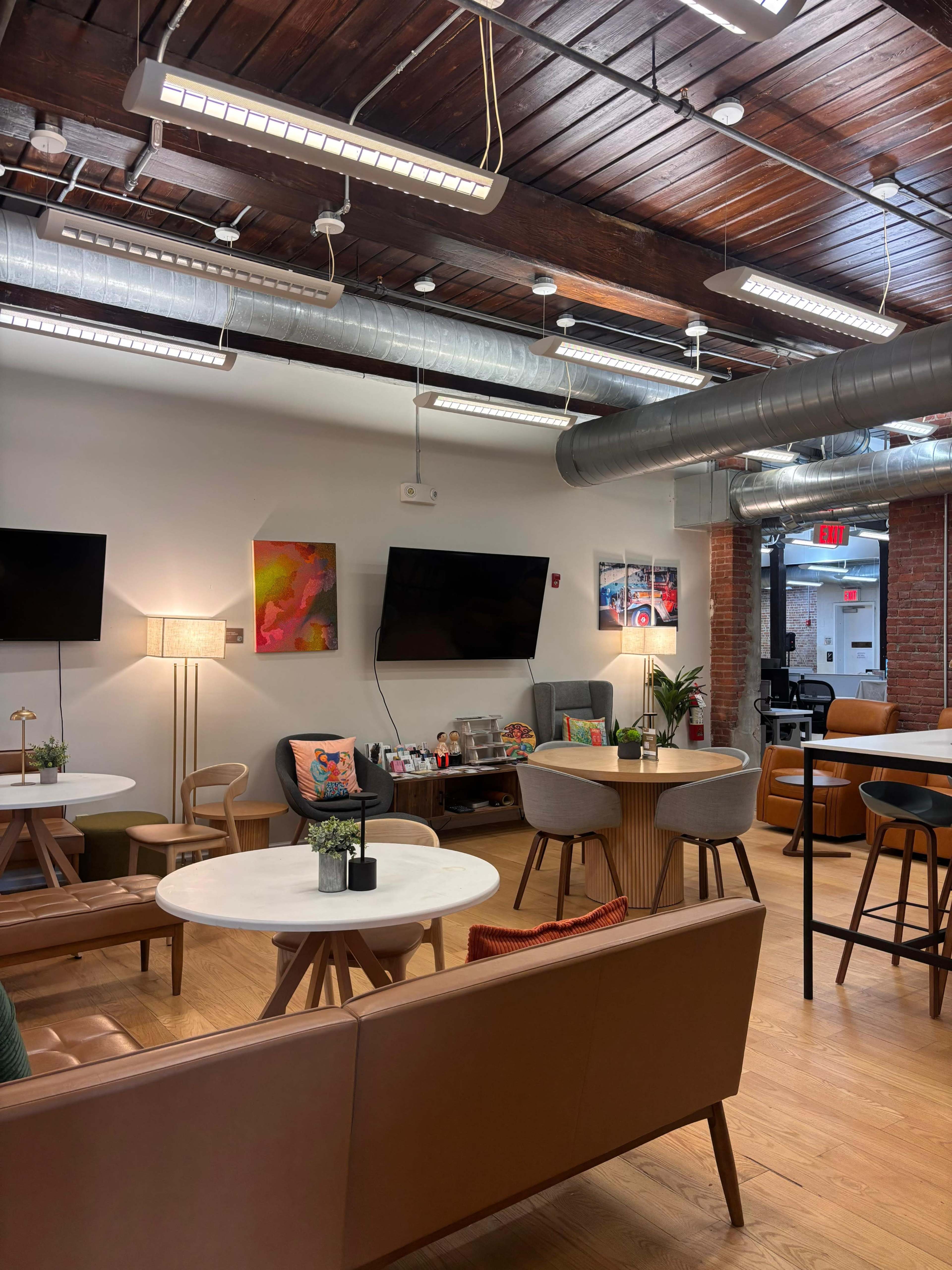 The image shows a modern lounge area with comfortable seating, two large screens, and decorative elements in a well-lit environment.