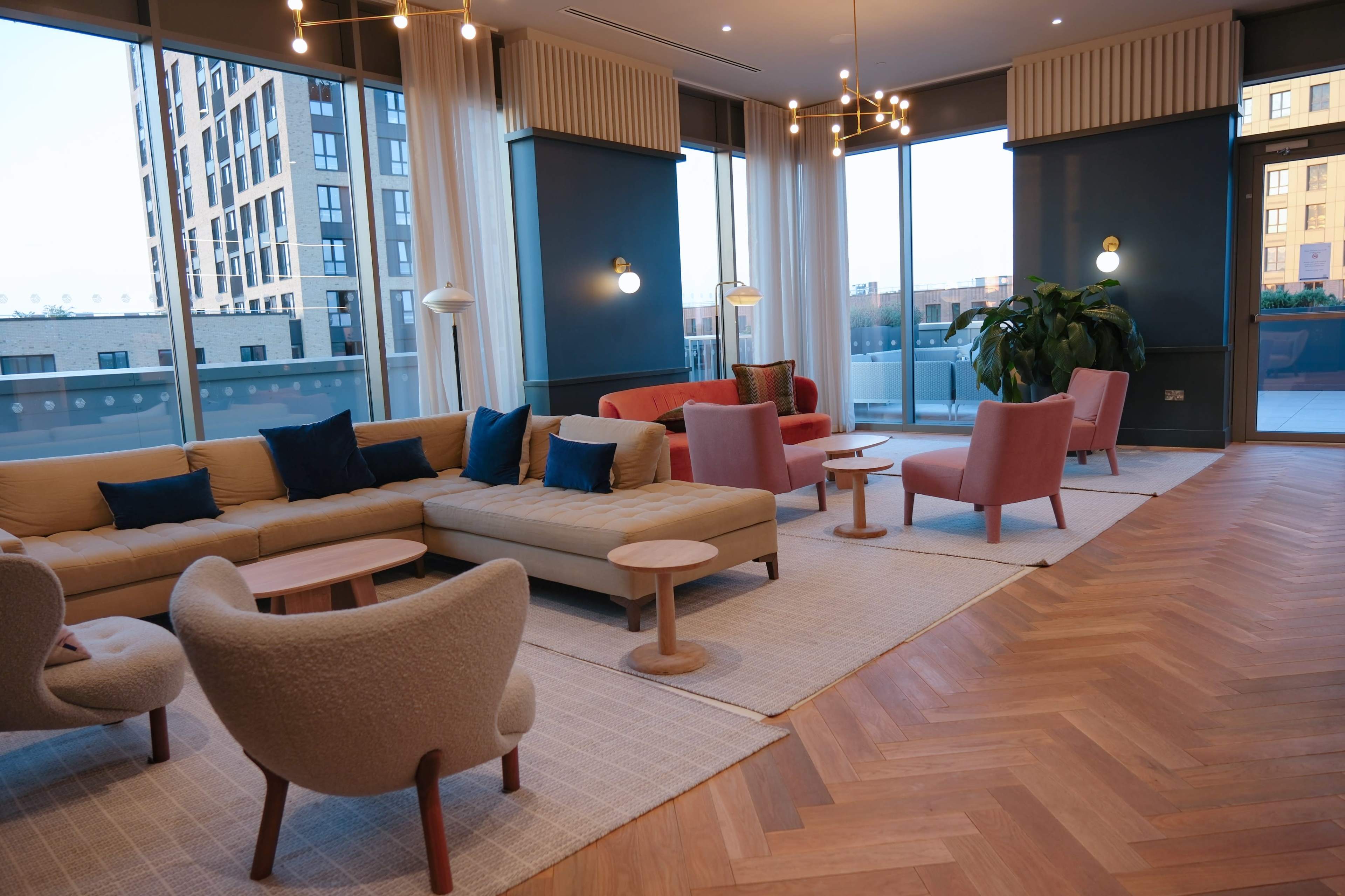 A modern lounge area featuring a large beige sofa, pink chairs, small wooden tables, and large windows that allow natural light to fill the space.