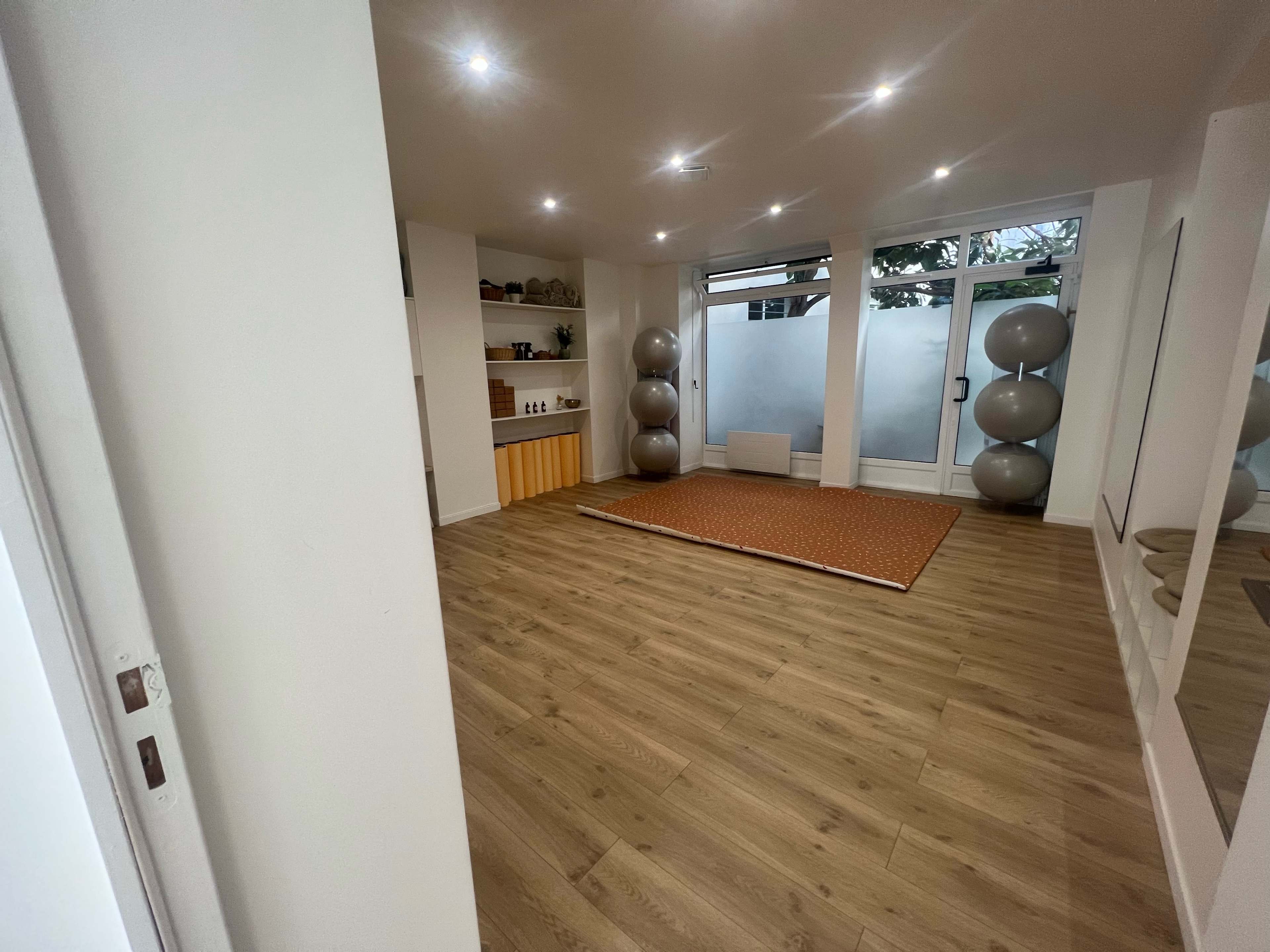 A spacious, well-lit room features wooden flooring, a large exercise mat, gray exercise balls, and a minimalist shelving unit against the wall.