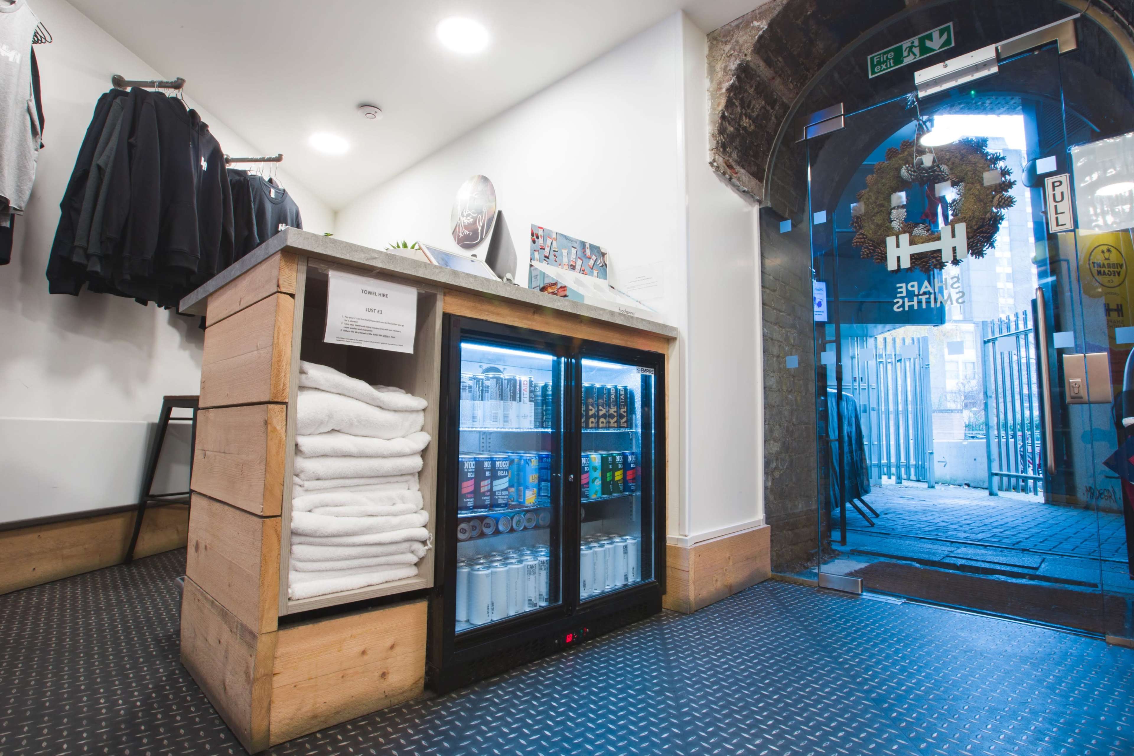 The image shows a neatly organized reception area with a wooden counter displaying a refrigerator stocked with drinks, towels stacked nearby, and an entrance leading to an outdoor area.
