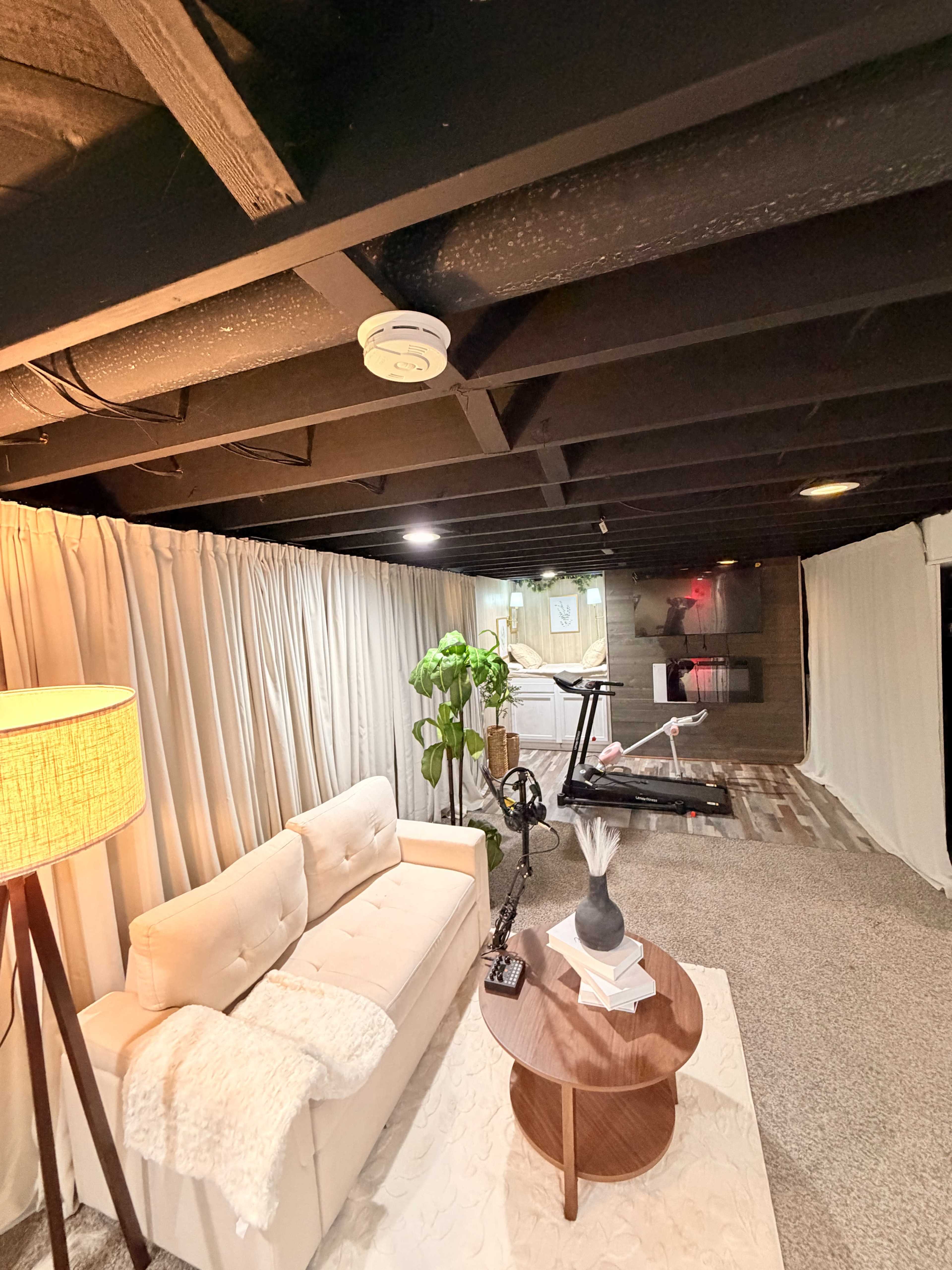 The image shows a cozy basement area with a sofa, a small table, and a potted plant, separated by curtains from another section.