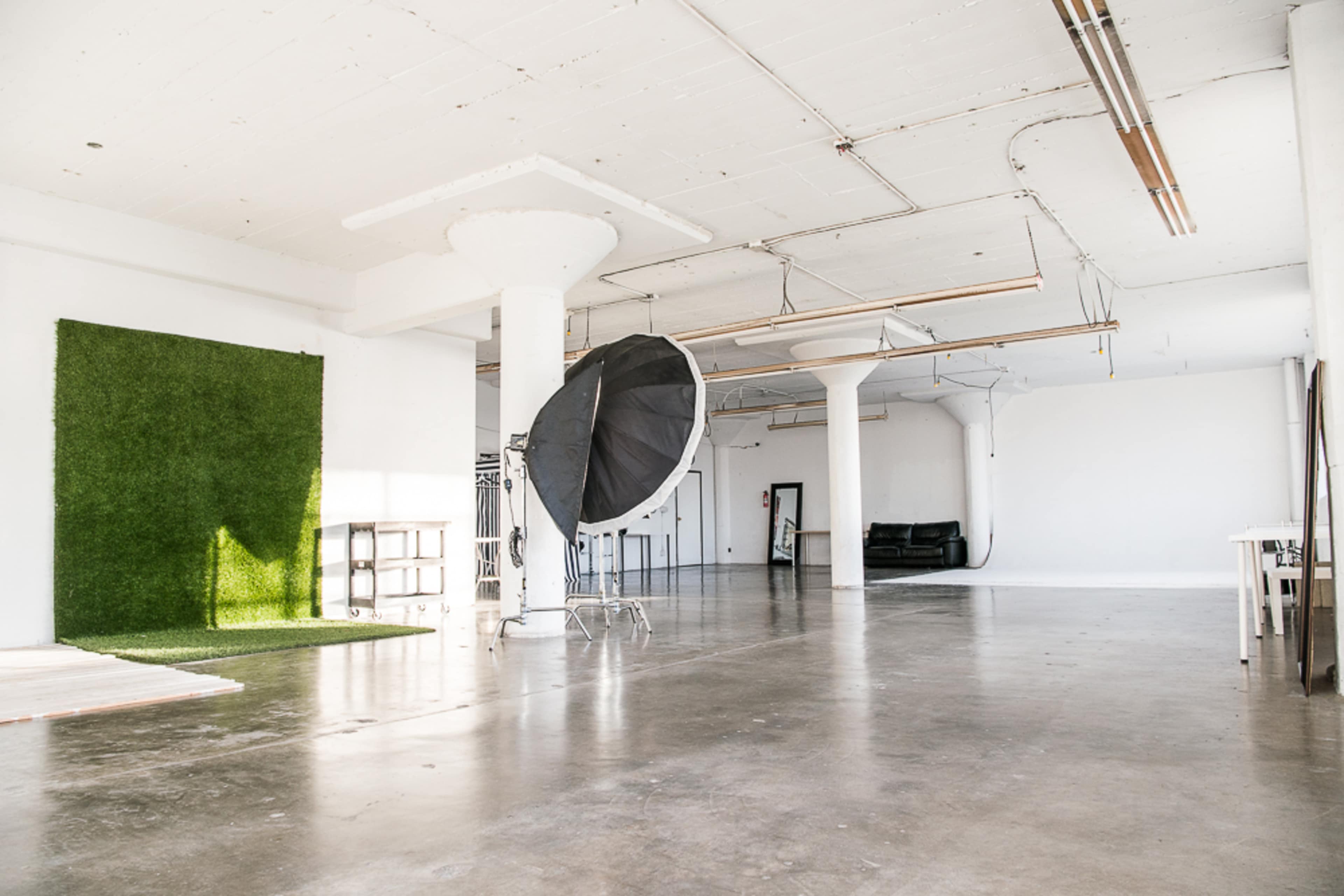 A spacious photo studio features a green backdrop, a large softbox, and minimal furniture against a white wall.