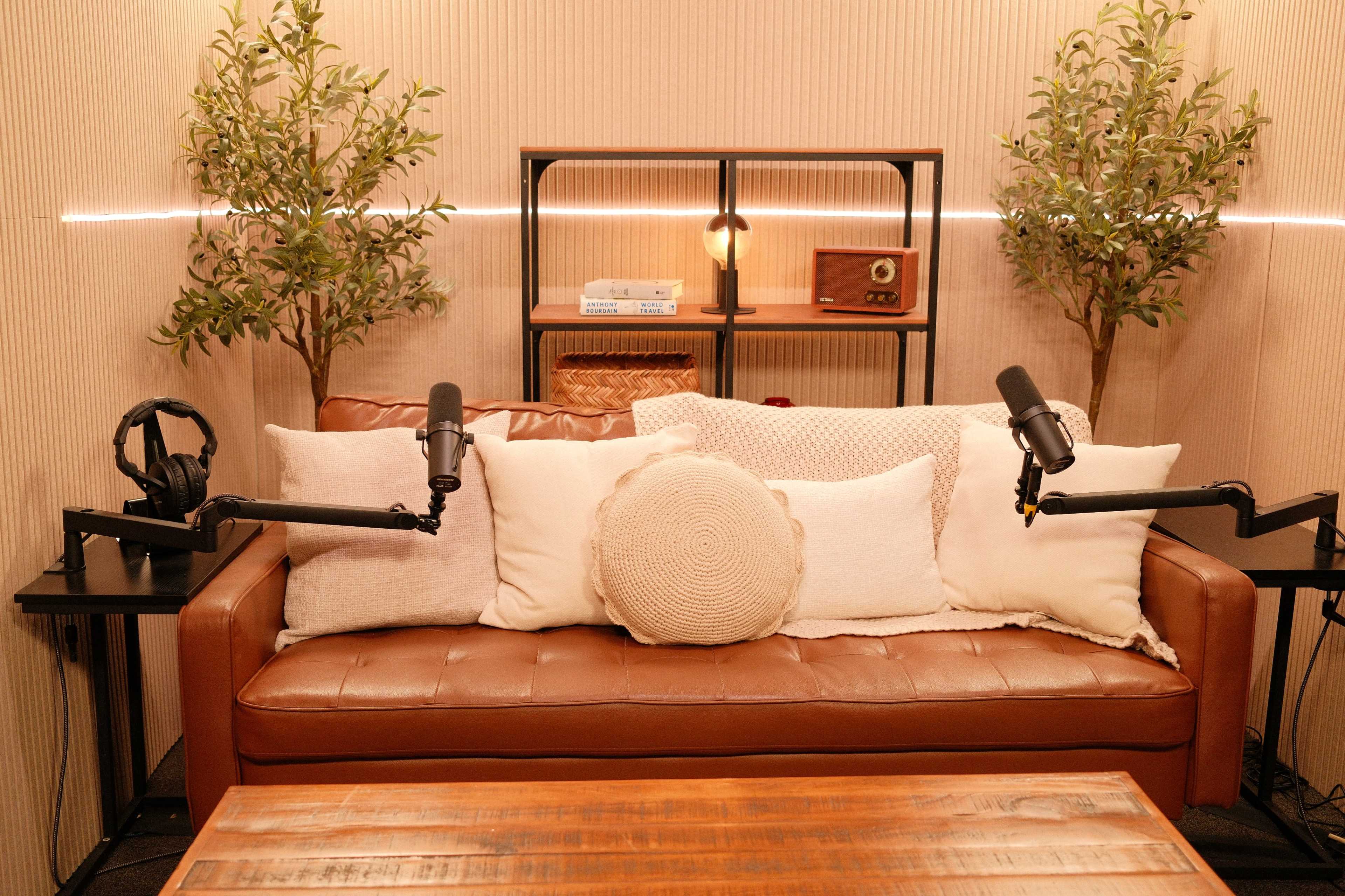 The image shows a cozy podcasting setup featuring a brown leather couch with several white pillows, two microphones mounted on arms, and small decorative trees on each side.