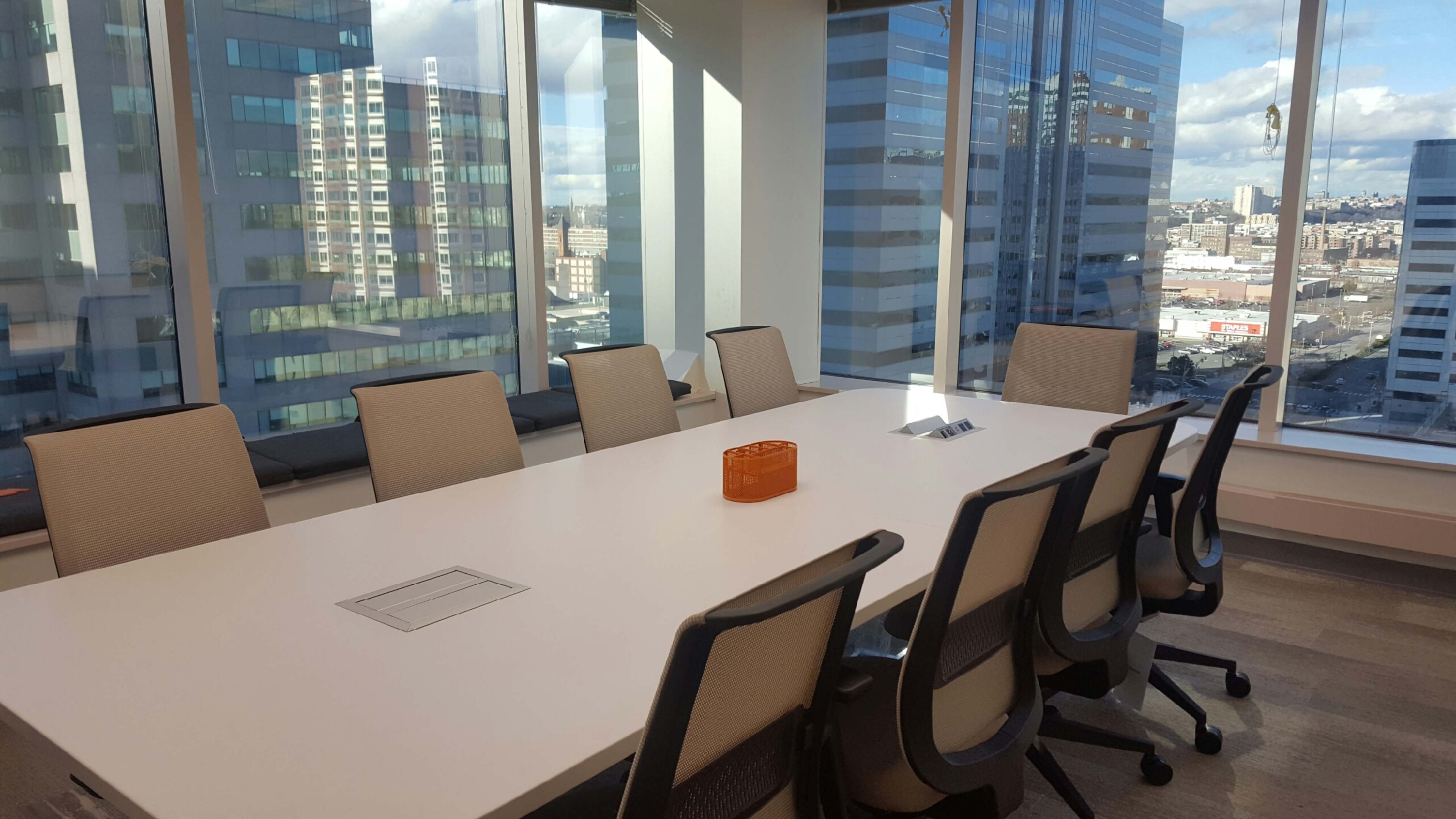 A large, modern conference room with a long white table and several ergonomic chairs, featuring windows that offer views of a city skyline.