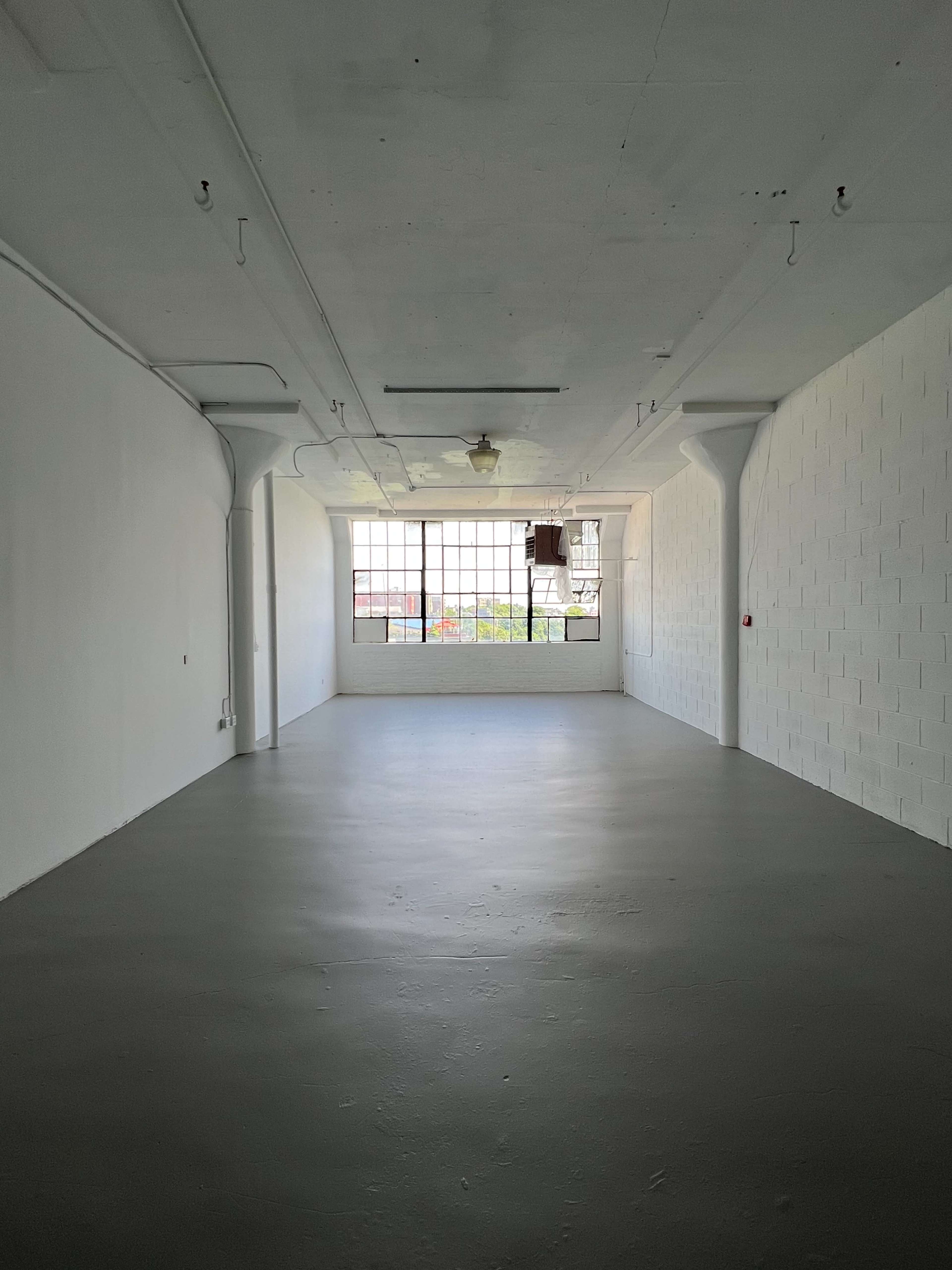 A spacious, empty room with white walls and a large window that allows natural light to enter.