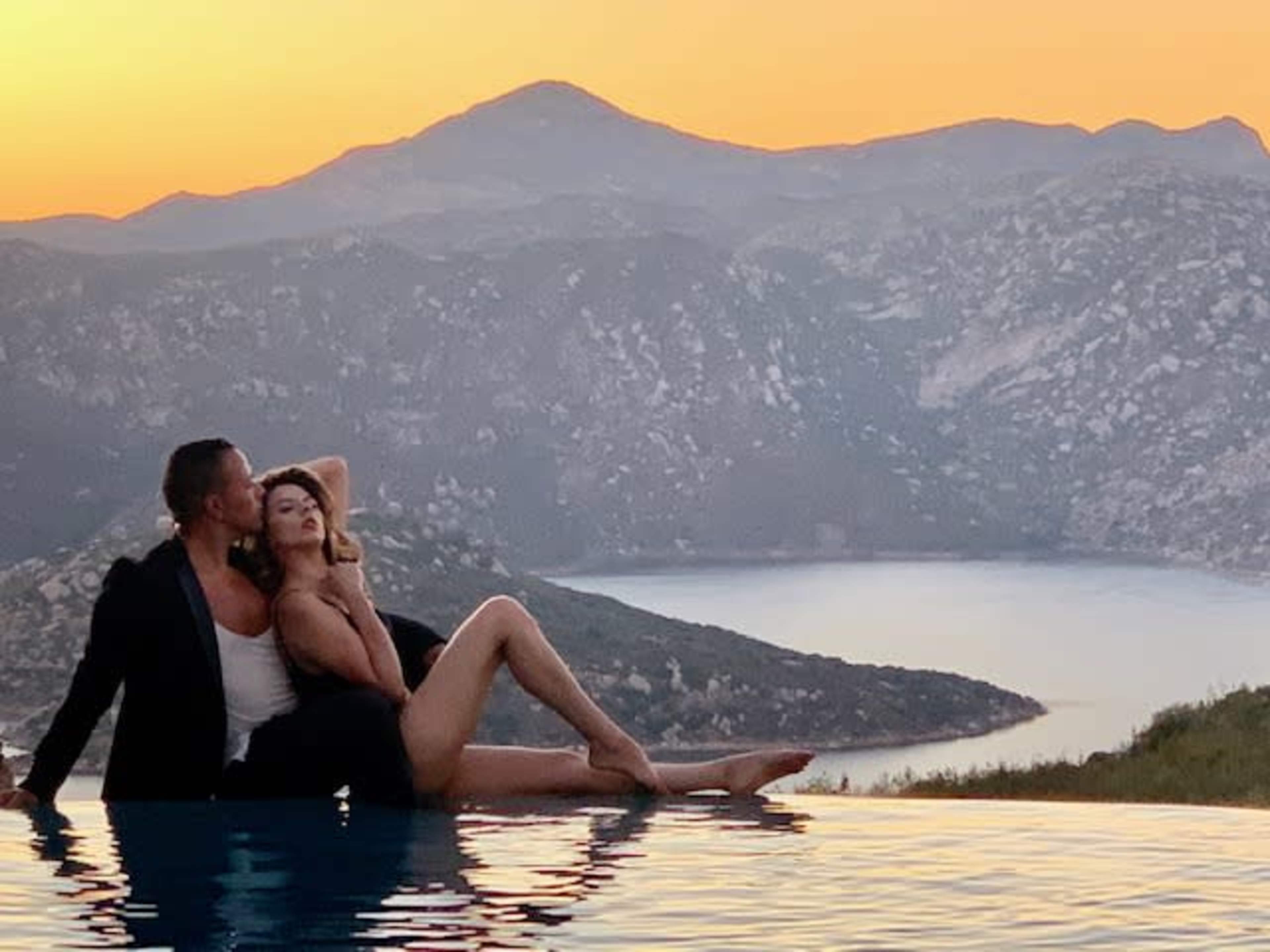 A couple poses affectionately by the edge of an infinity pool overlooking a mountainous landscape at sunset.