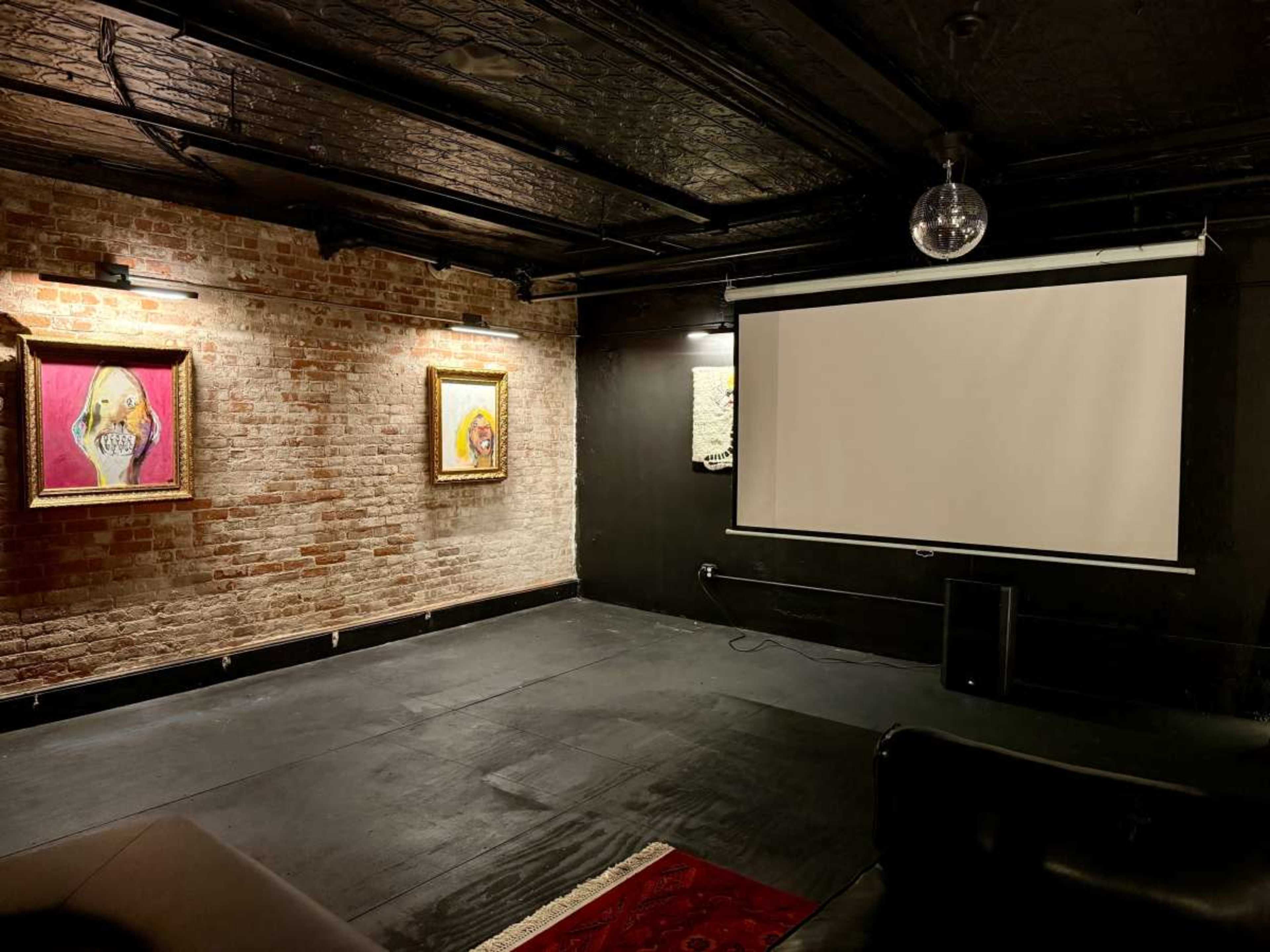 The image shows a dimly lit art space with exposed brick walls, framed paintings, and a blank projection screen.