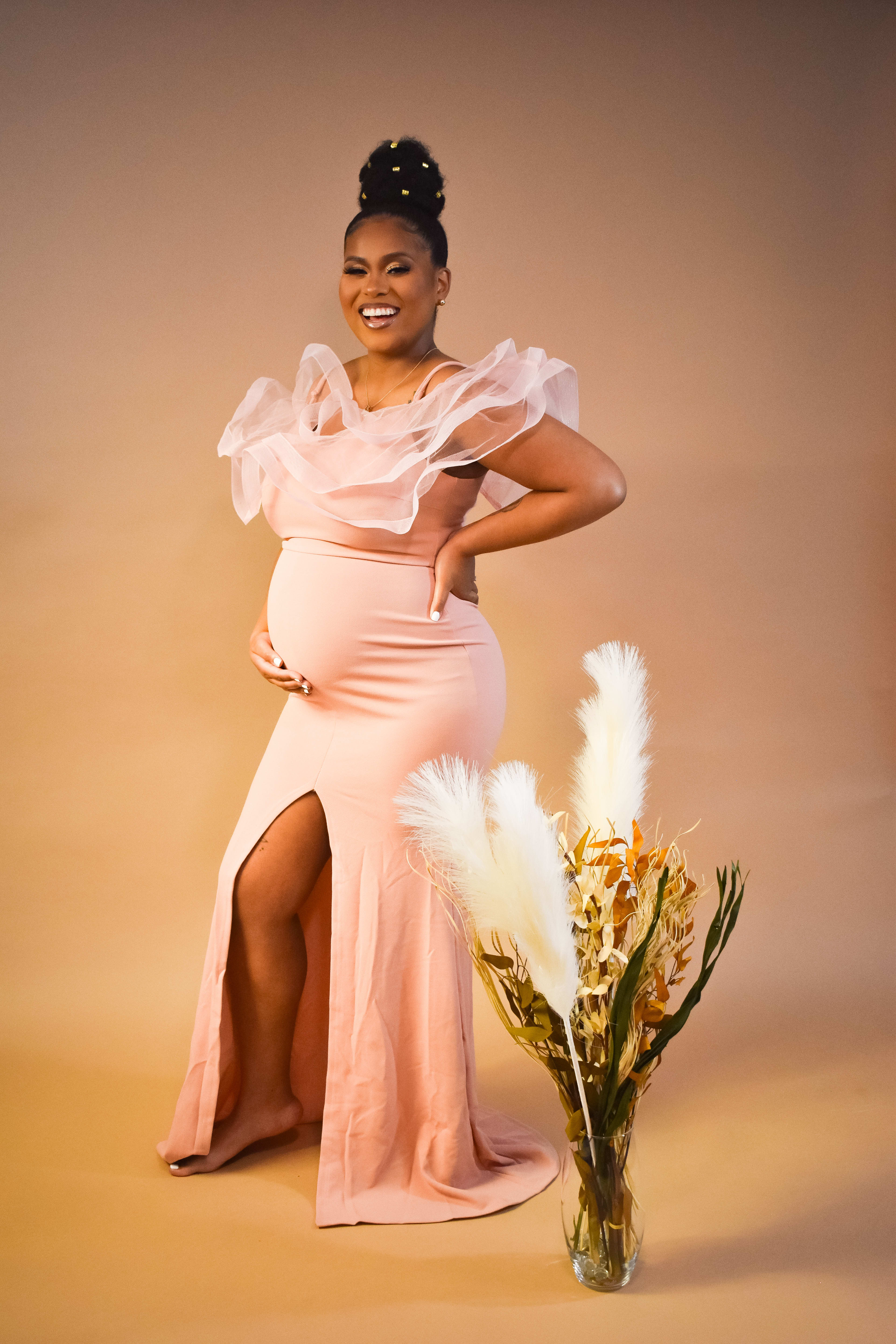 A pregnant woman in a pink boho dress posing next to a vase of flowers during a maternity photo shoot.