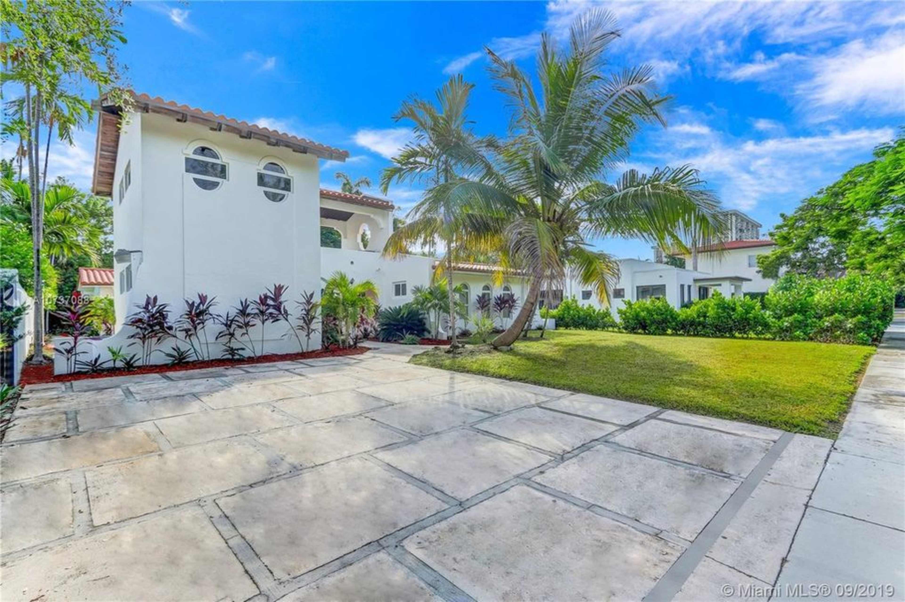 A sunny residential property featuring a white house with round windows, palm trees, and a paved driveway surrounded by greenery.