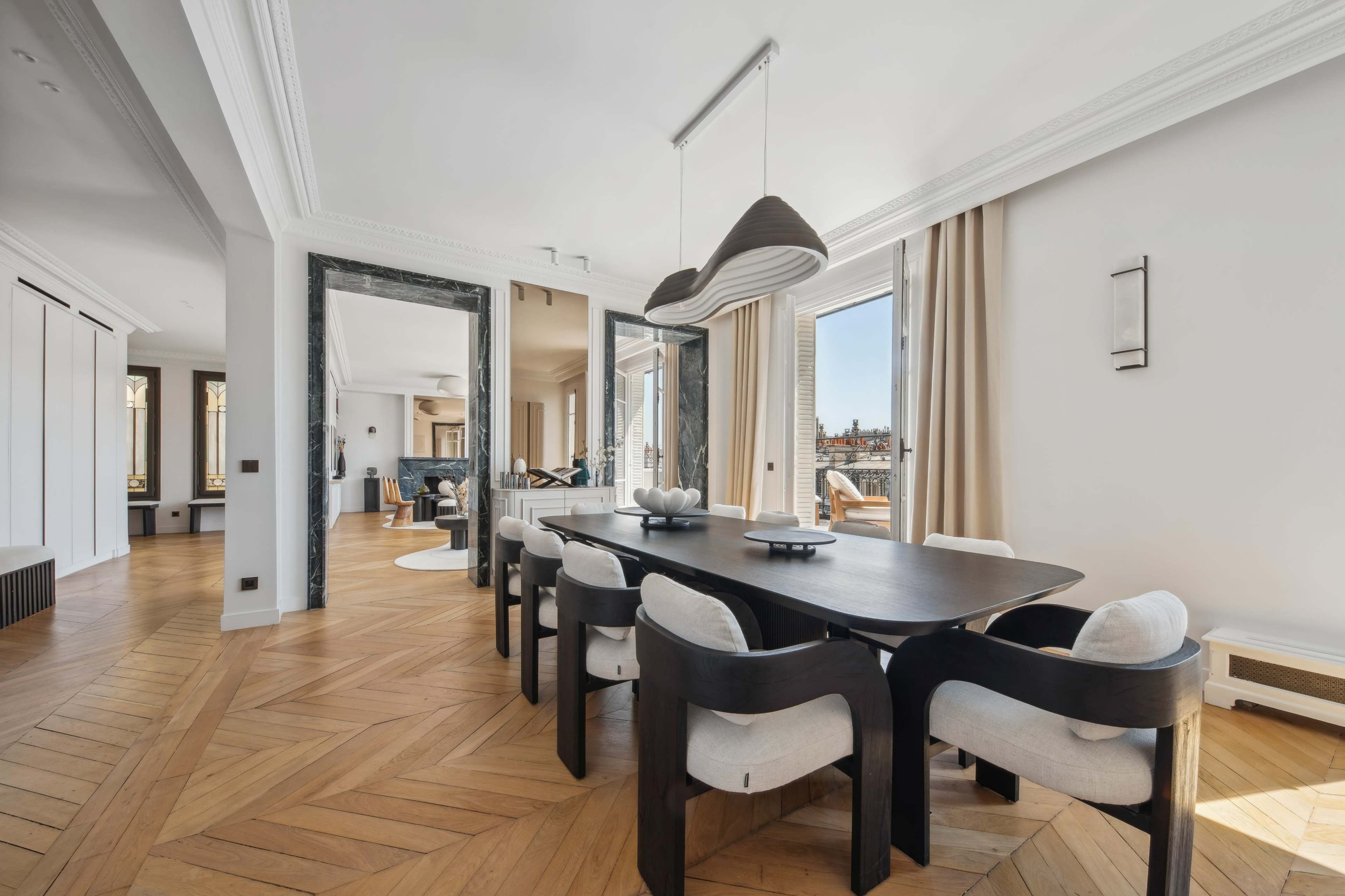 A dining room features a dark wooden table surrounded by upholstered chairs, with large windows offering views of the city and mirrored walls enhancing the space.