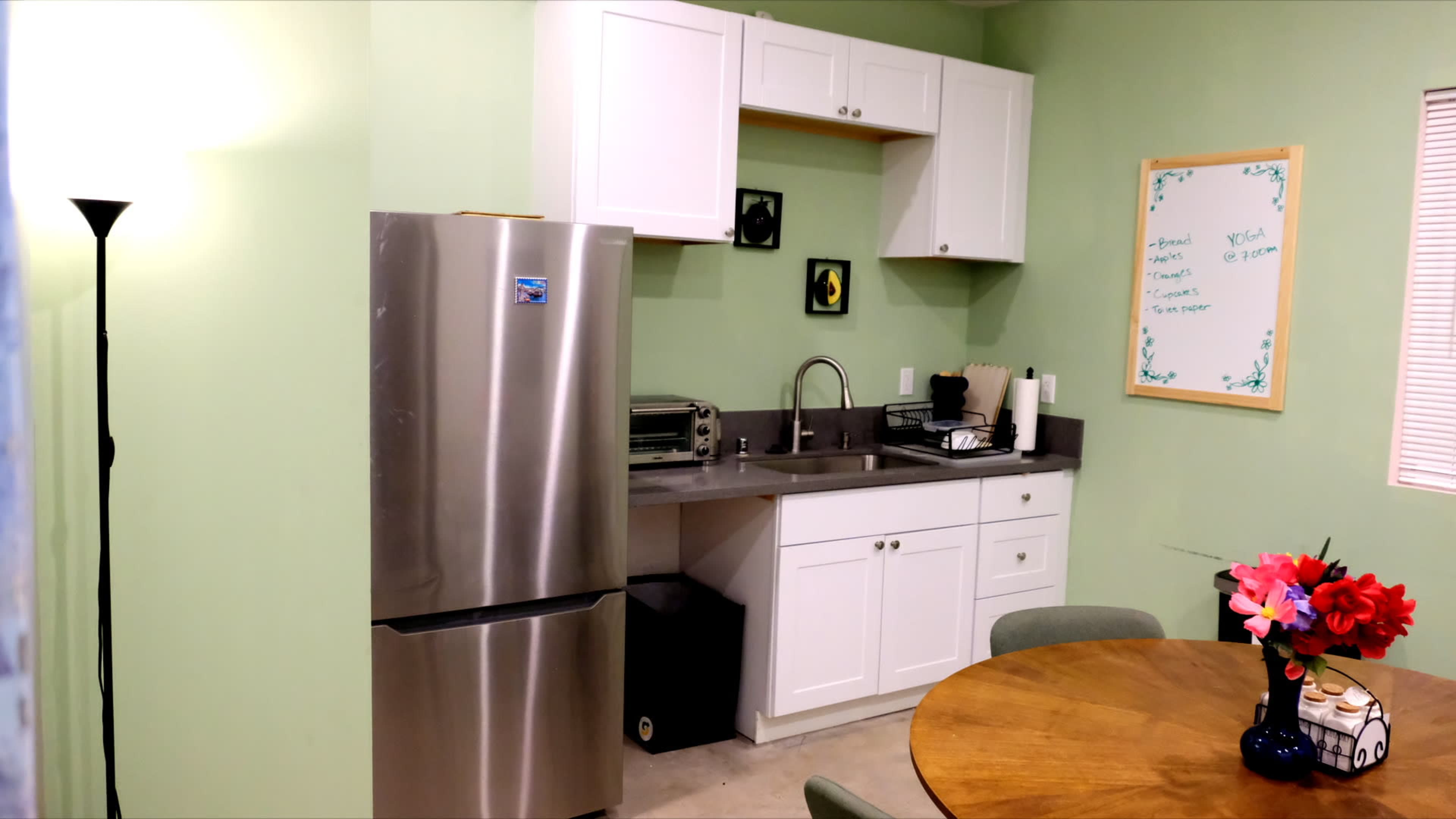 The image shows a small kitchen area with green walls, featuring stainless steel appliances, a wooden dining table with flowers, and a whiteboard on the wall.