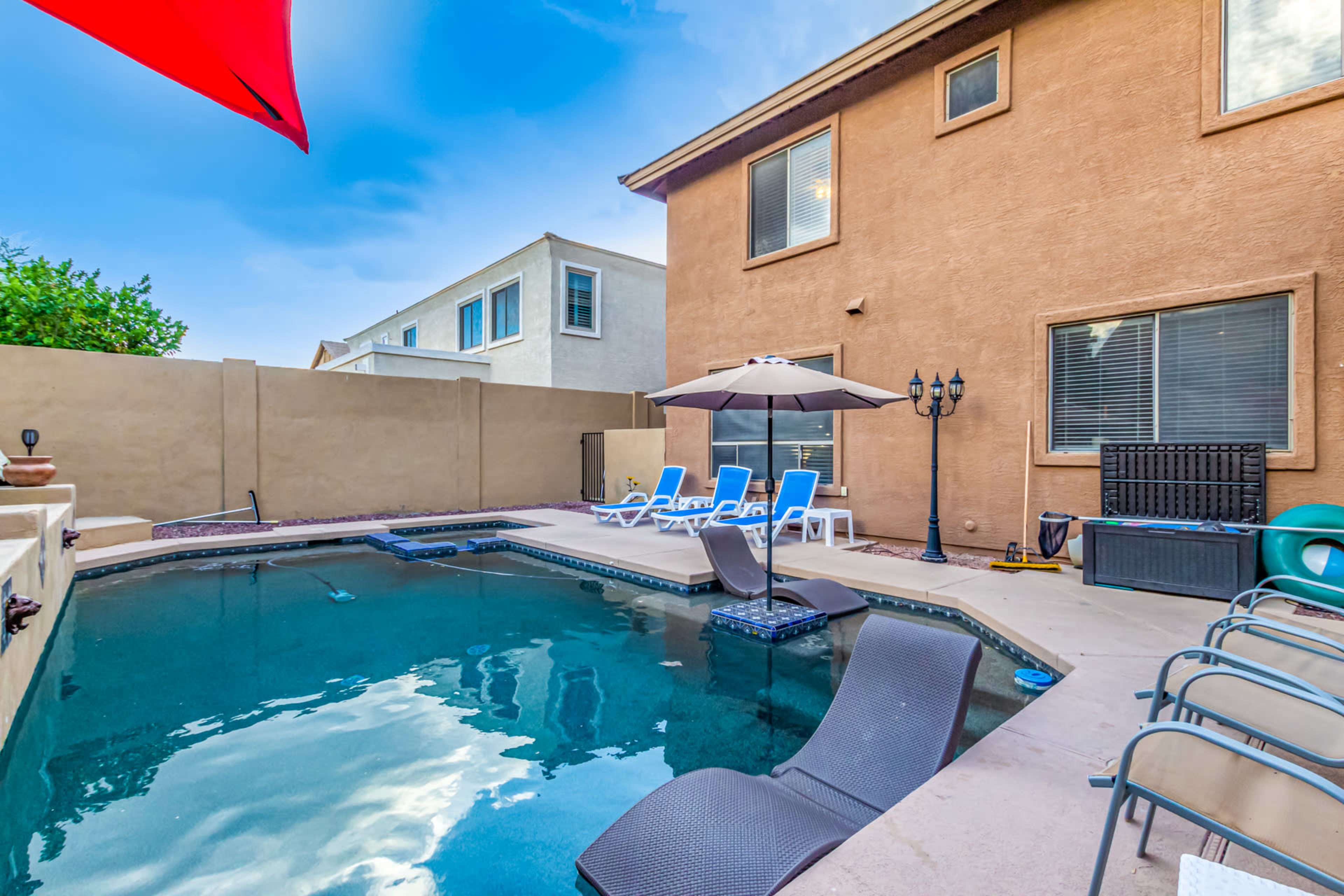 A swimming pool area features lounge chairs and an umbrella, surrounded by a beige wall and a two-story building.