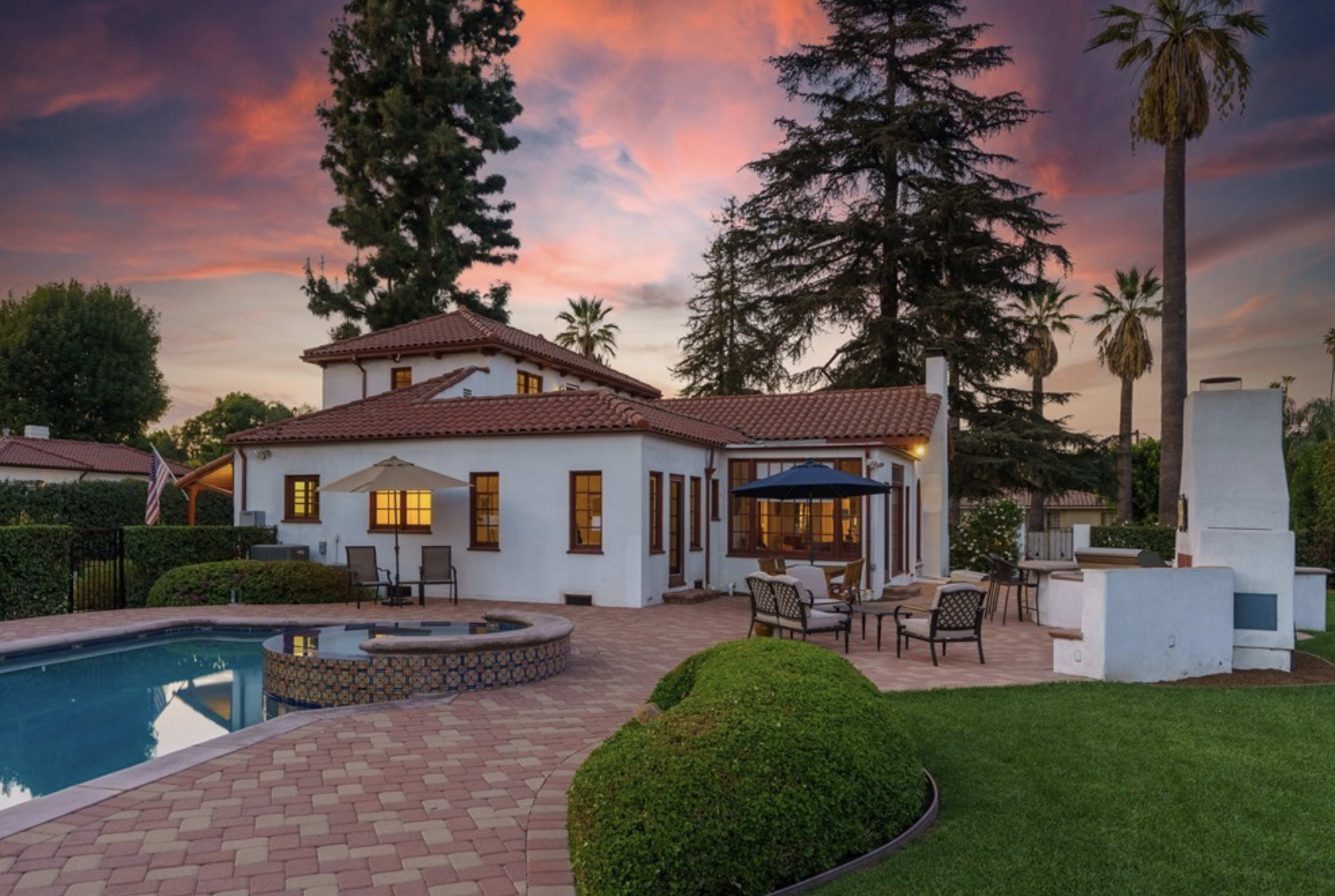 A two-story house with a tiled roof, surrounded by a well-maintained yard, a swimming pool, and palm trees at sunset.