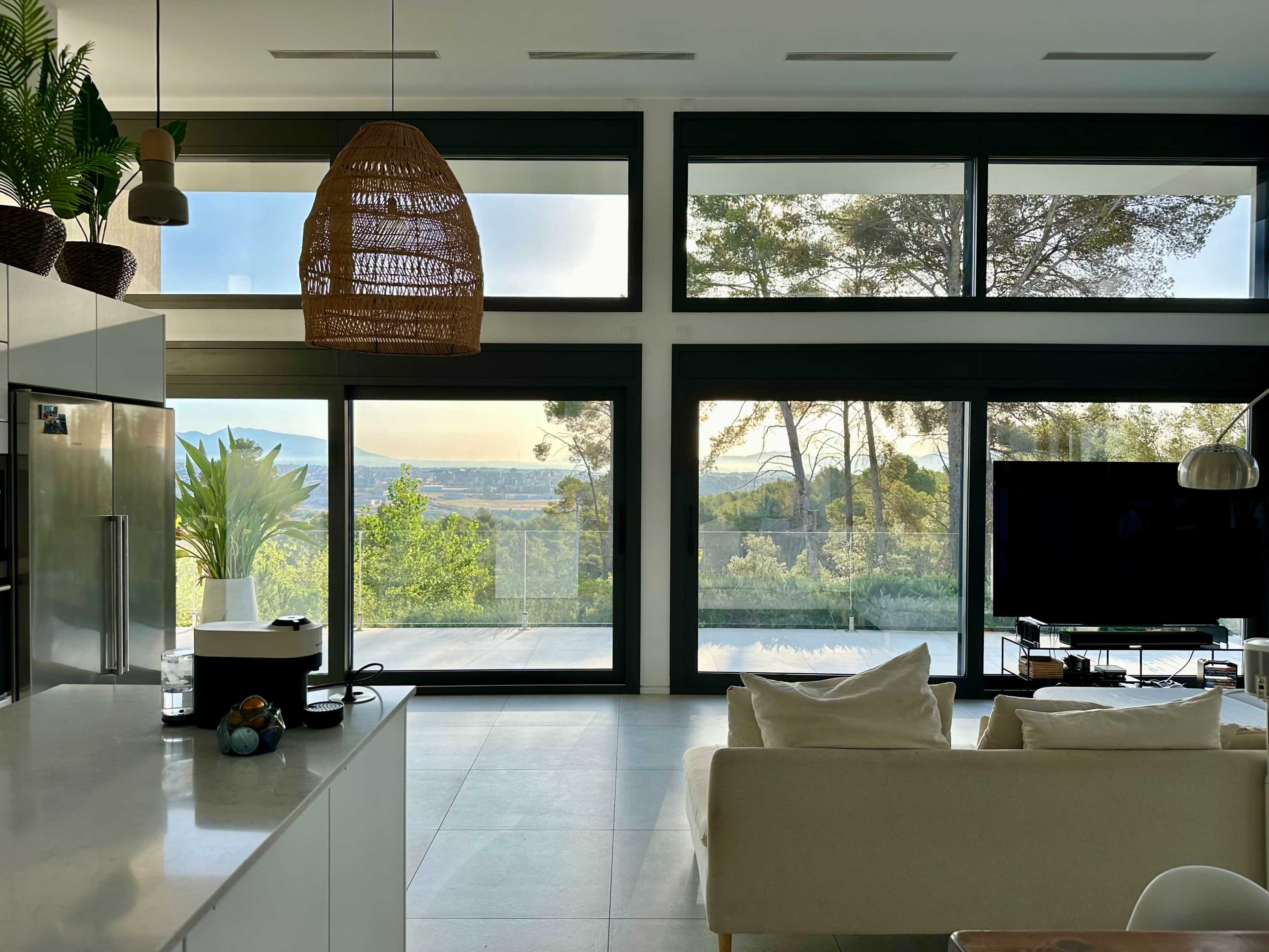A modern living room features large glass windows with a view of trees and distant hills, furnished with a cream couch and a contemporary light fixture.