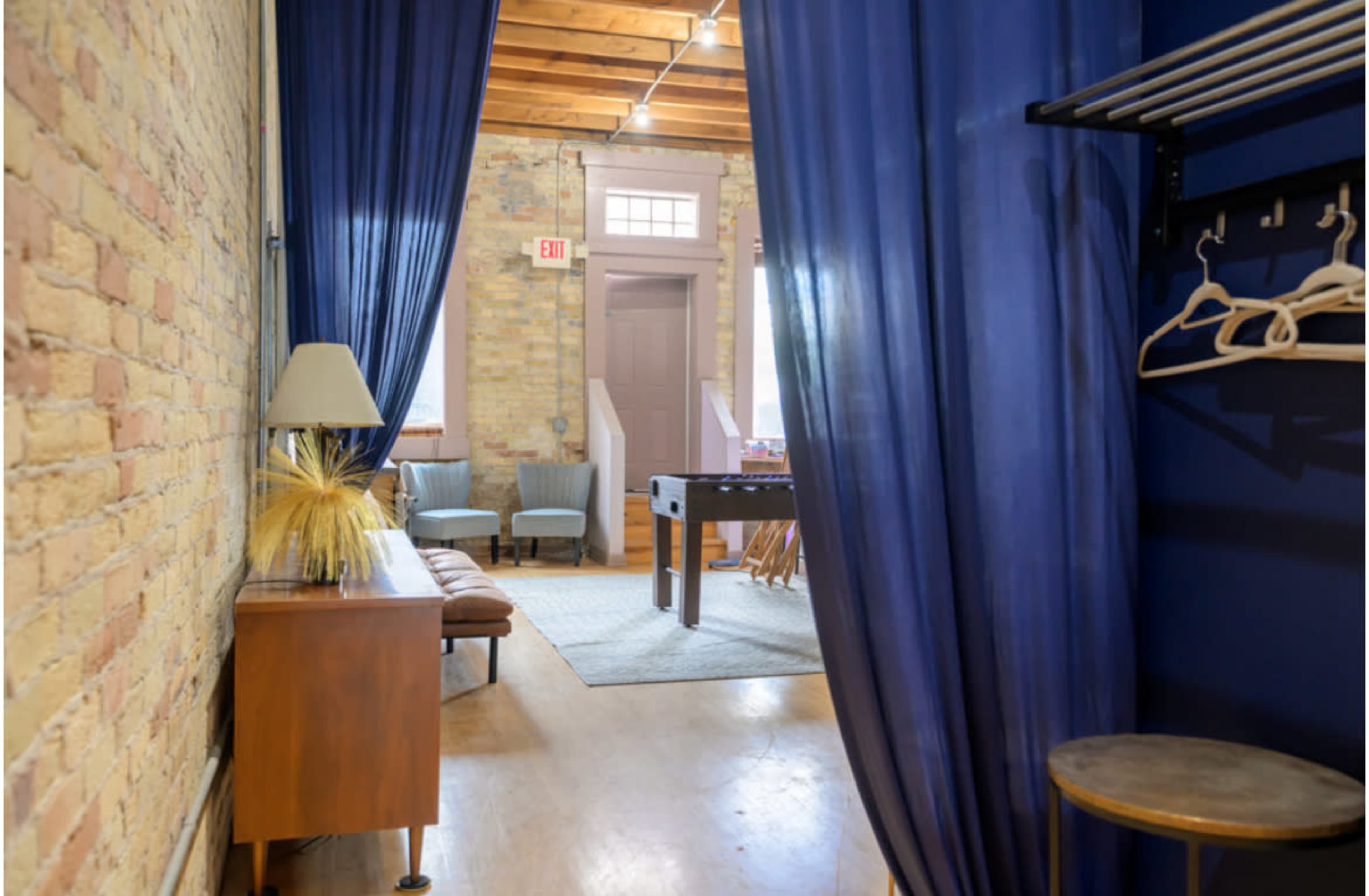 The image shows a hallway leading to a brightly lit room with blue curtains, featuring a seating area and a foosball table in the background.