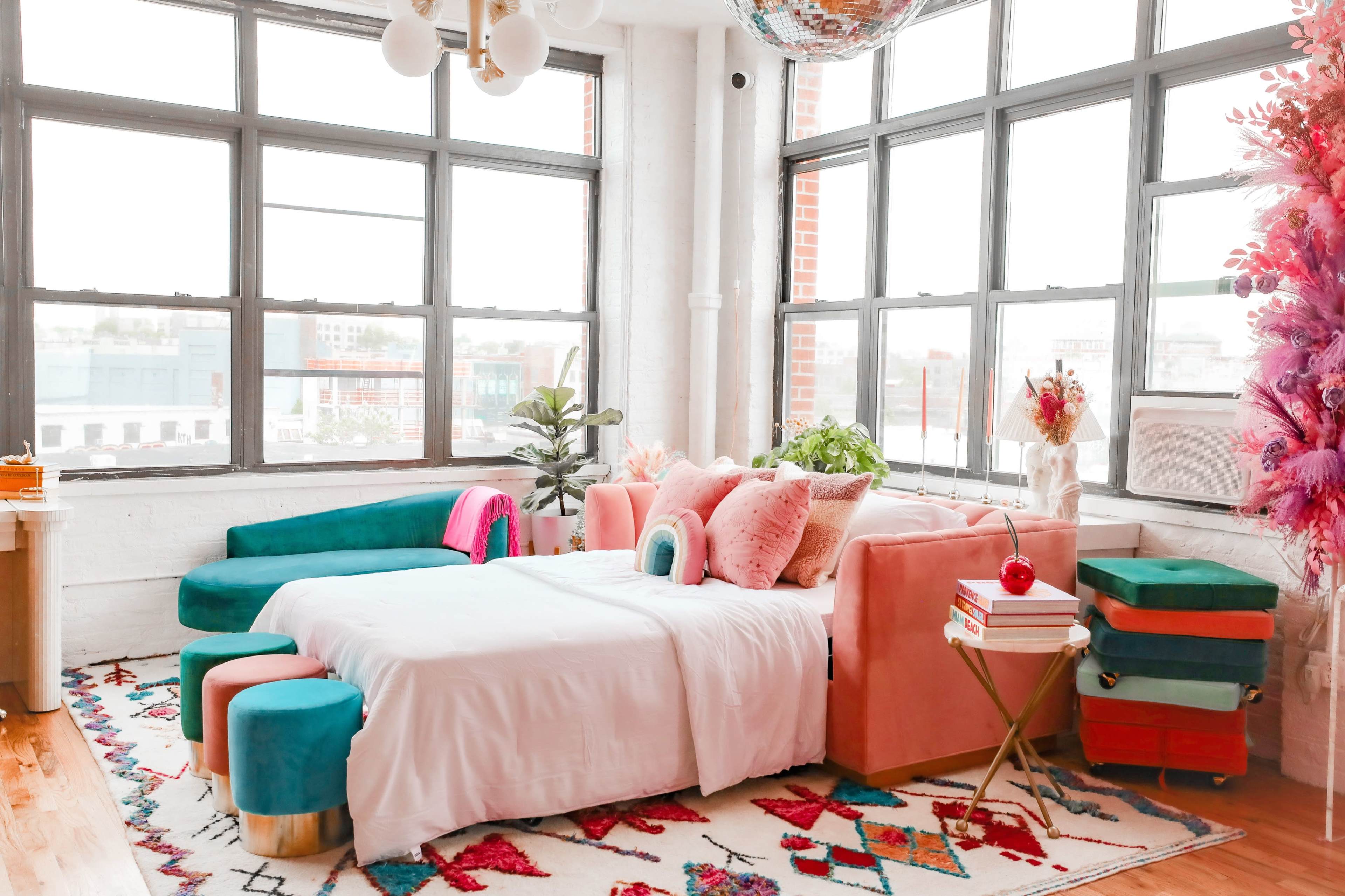 A brightly lit room features a pink sofa with decorative pillows, a vibrant rug, and large windows offering an urban view.