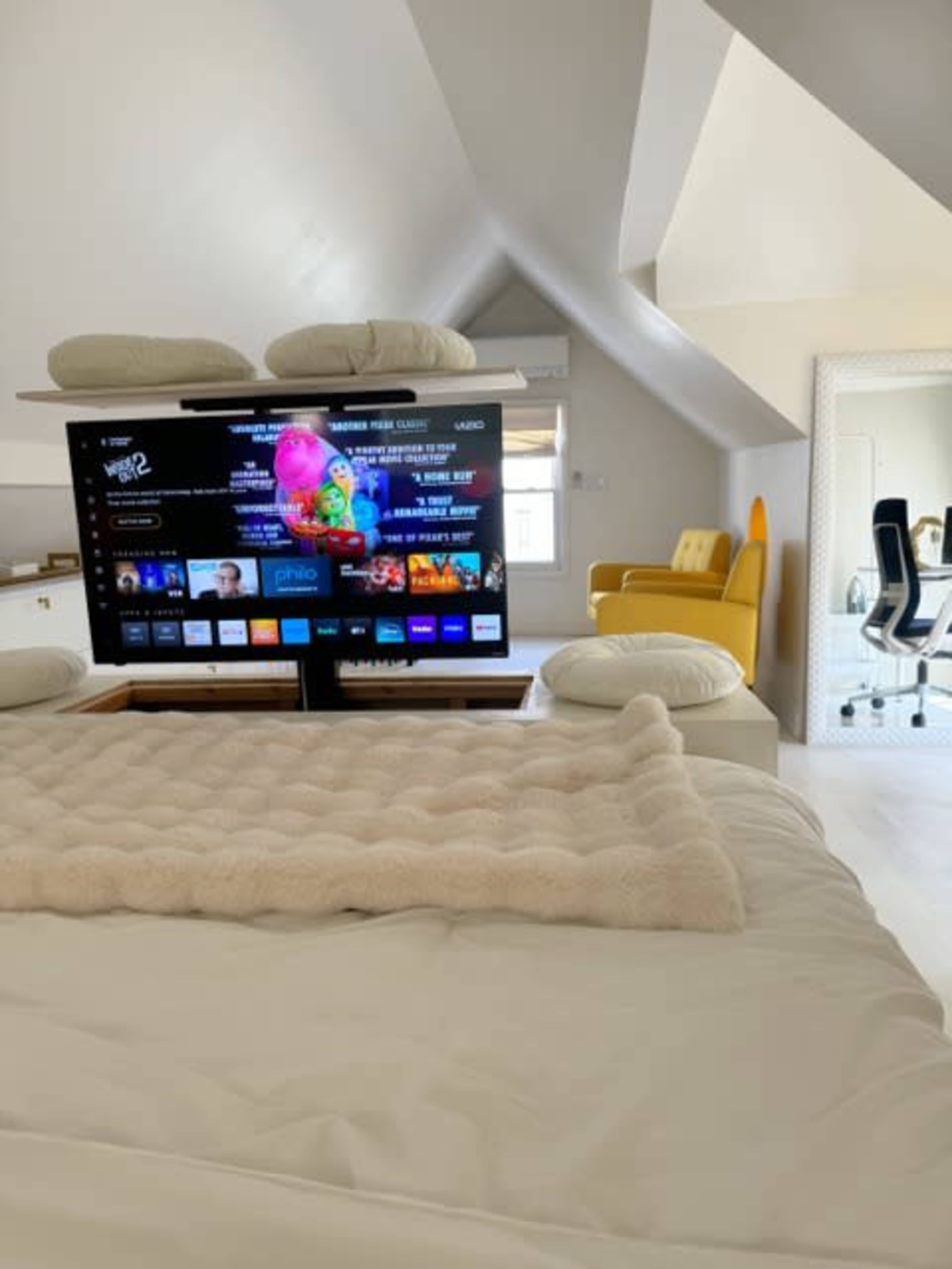 The image shows a cozy bedroom with a large TV mounted on a shelf, a bed with white bedding, and a yellow chair in the background.