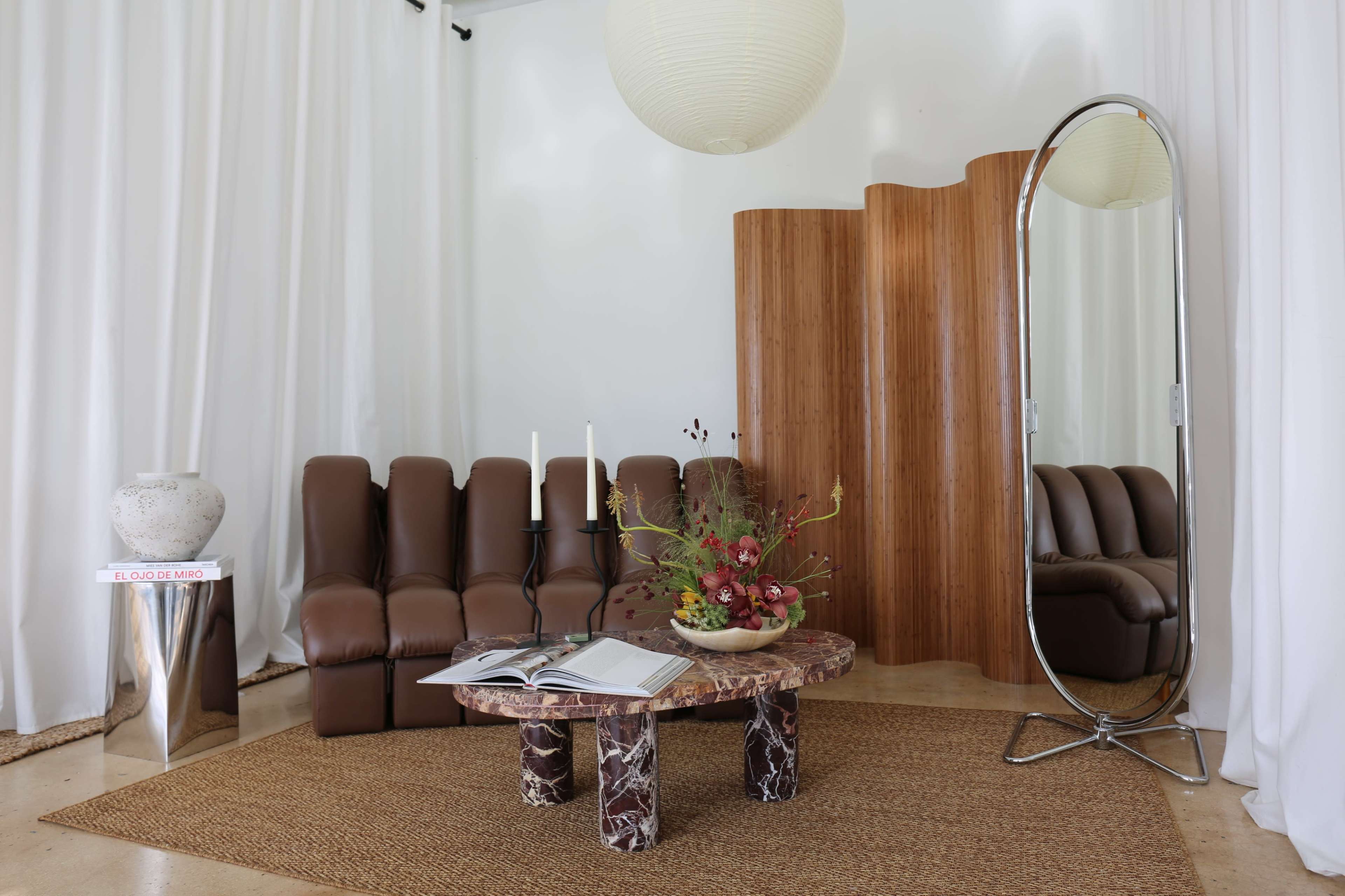 The scene features a modern living room with a brown leather sofa, a marble coffee table, a large mirror, and a decorative arrangement of flowers.