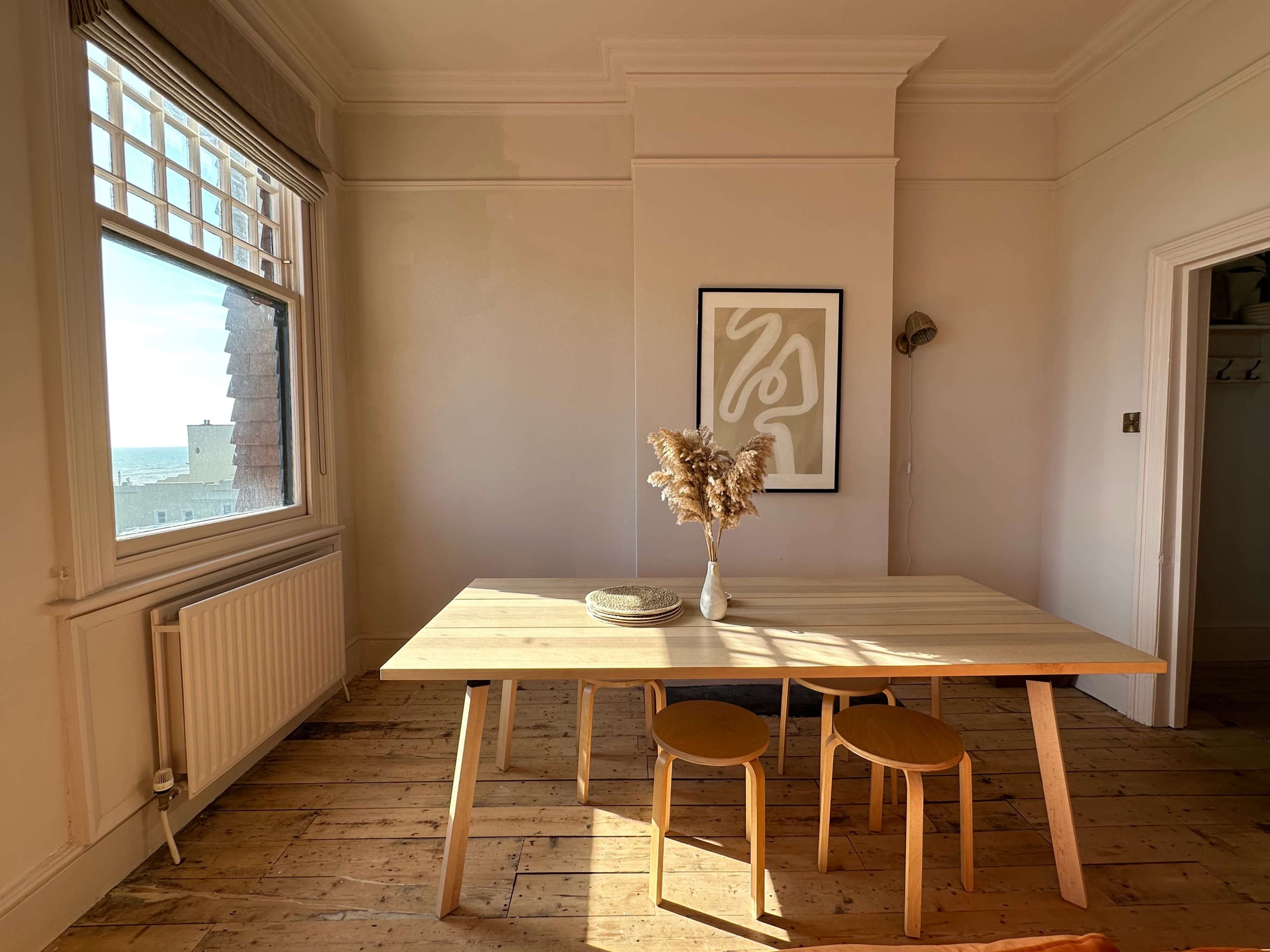 A wooden dining table with two chairs is positioned near a window, adorned with a vase of dried flowers and a decorative plate.