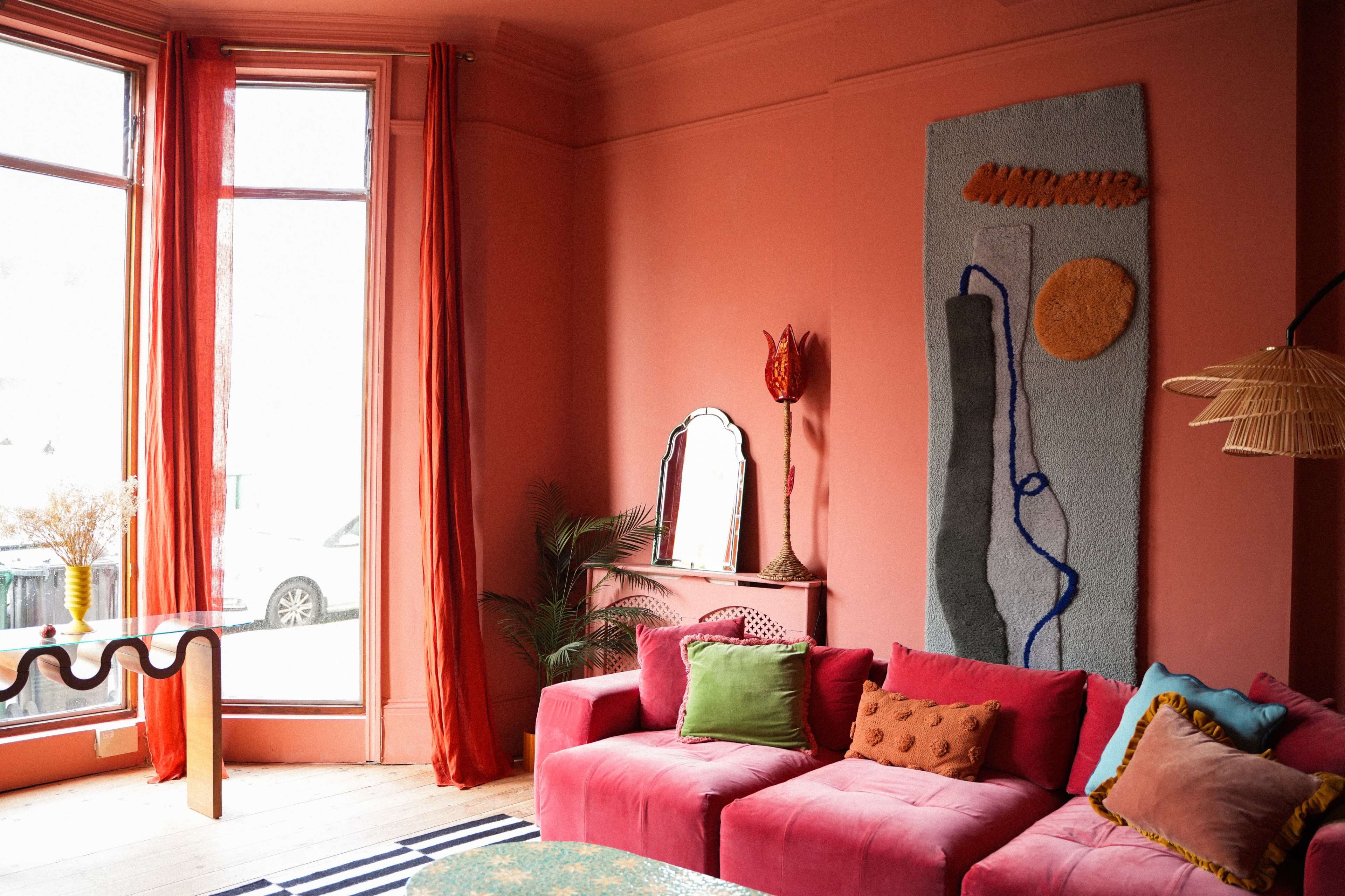 A bright living room features a pink wall, a sectional couch with colorful cushions, a decorative textile hanging, and a large window with sheer curtains.