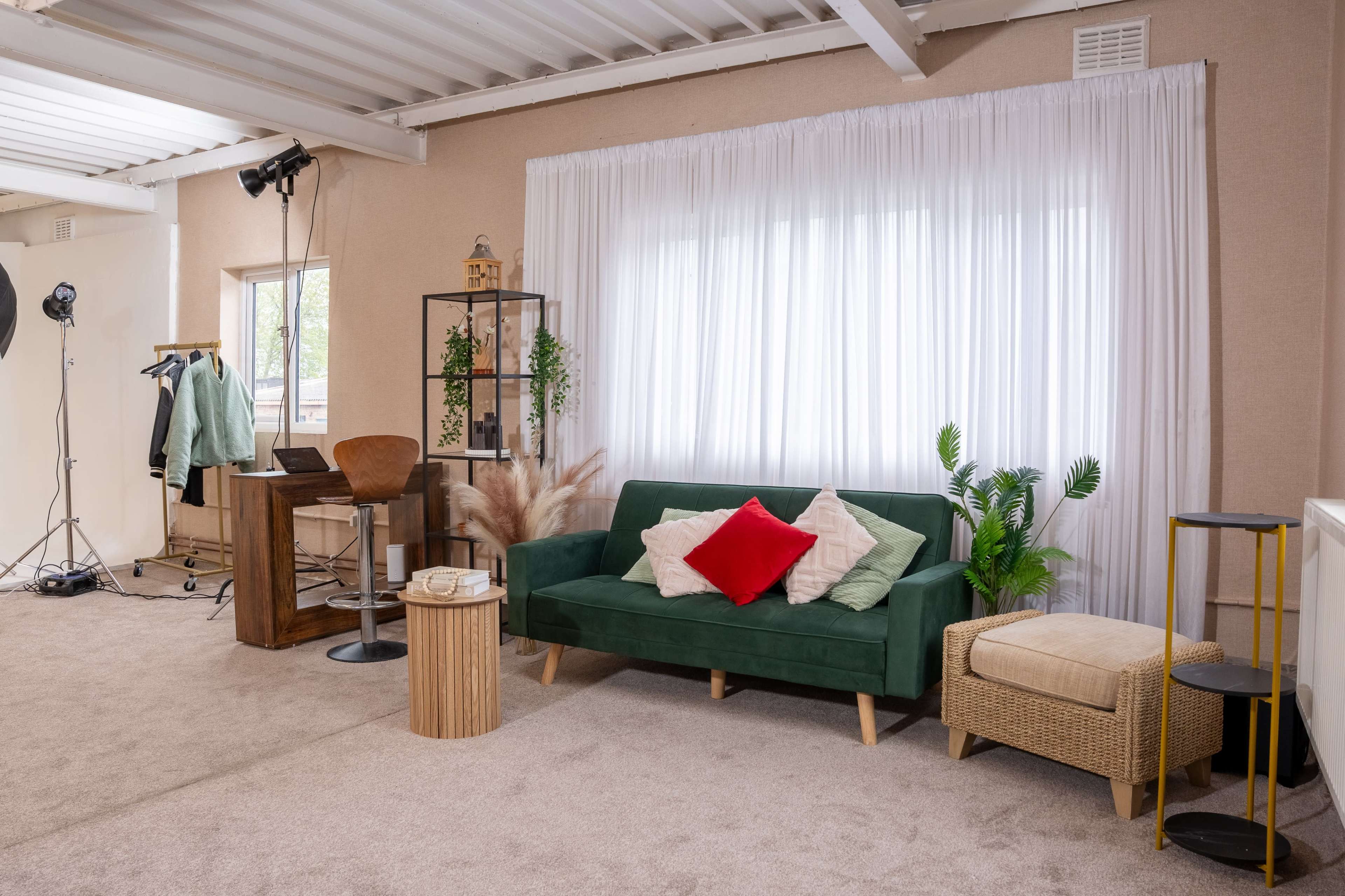 The scene features a cozy living space with a green sofa adorned with red and white throw pillows, a small wooden table, and a desk with a chair, all arranged near a large window covered by white curtains.