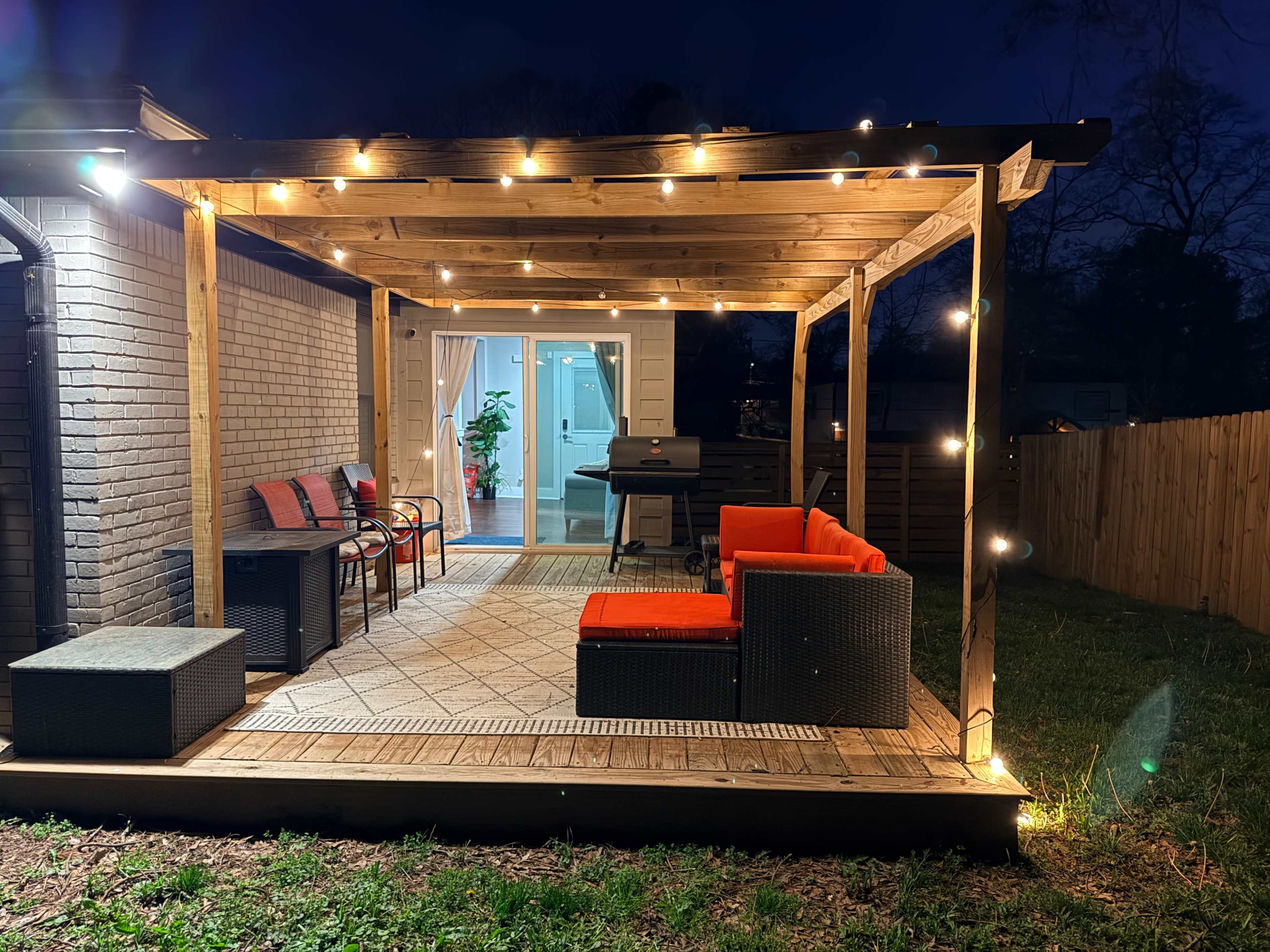 The image shows a well-lit patio with a wooden pergola, featuring seating furniture and a barbecue grill, set against a dark evening sky.