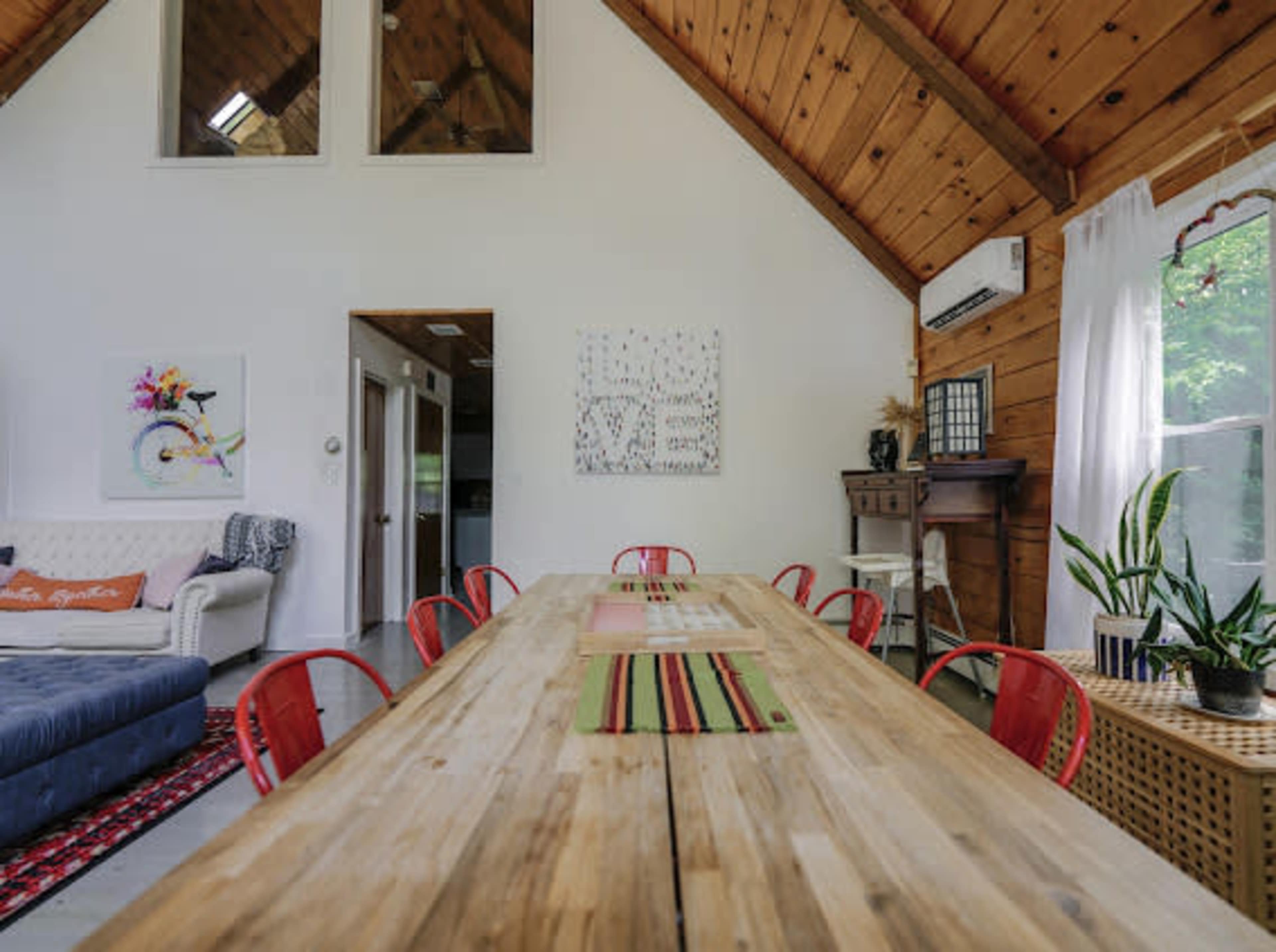 A spacious dining area features a long wooden table surrounded by red chairs, with a cozy couch and large windows in the background.