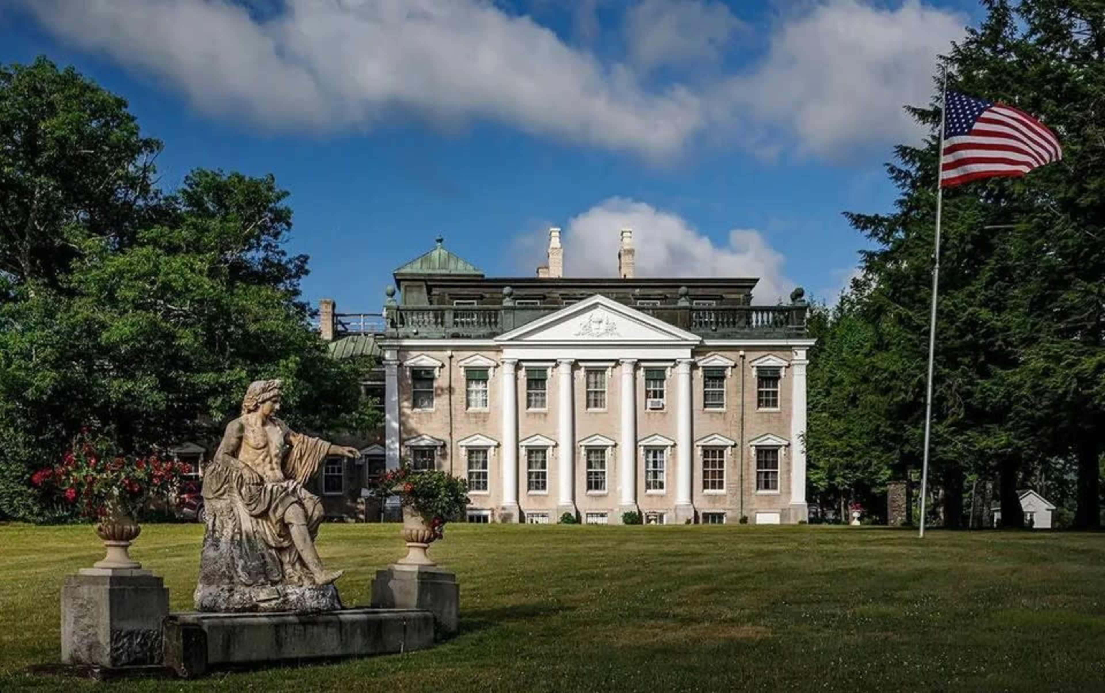 A large neoclassical mansion with columns stands behind a statue and an American flag, surrounded by green lawns and trees under a partly cloudy sky.