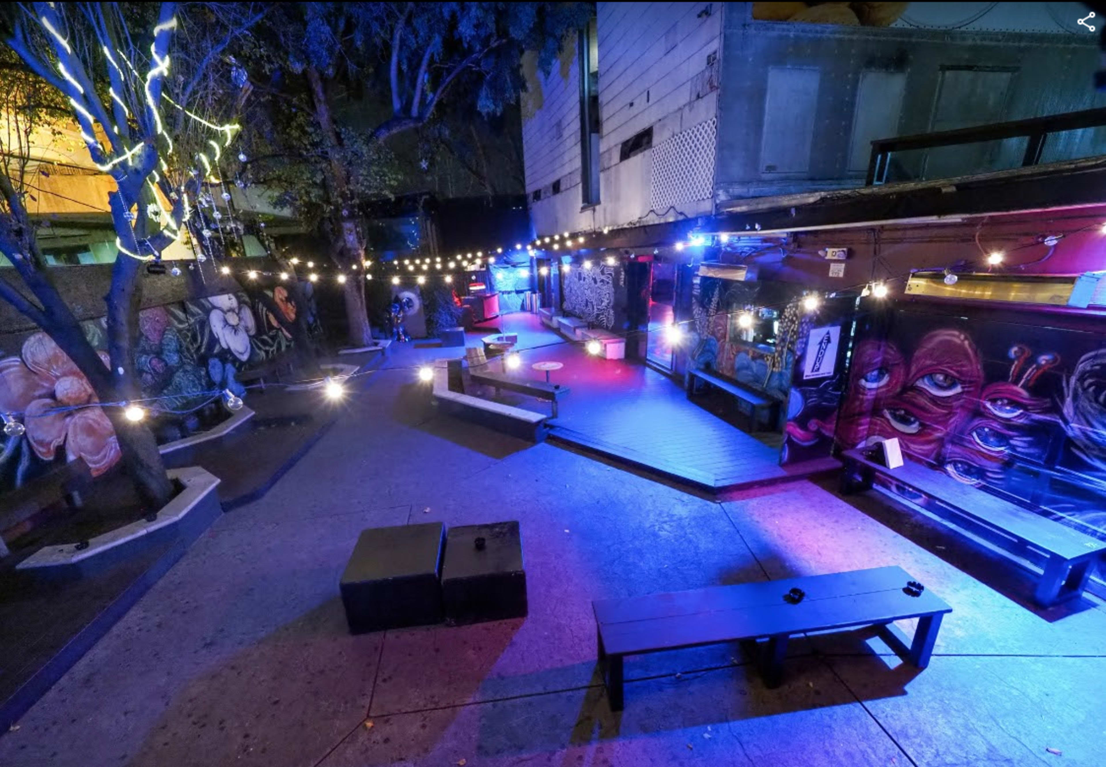An outdoor bar area with string lights illuminating colorful murals on the walls and wooden seating arrangements.