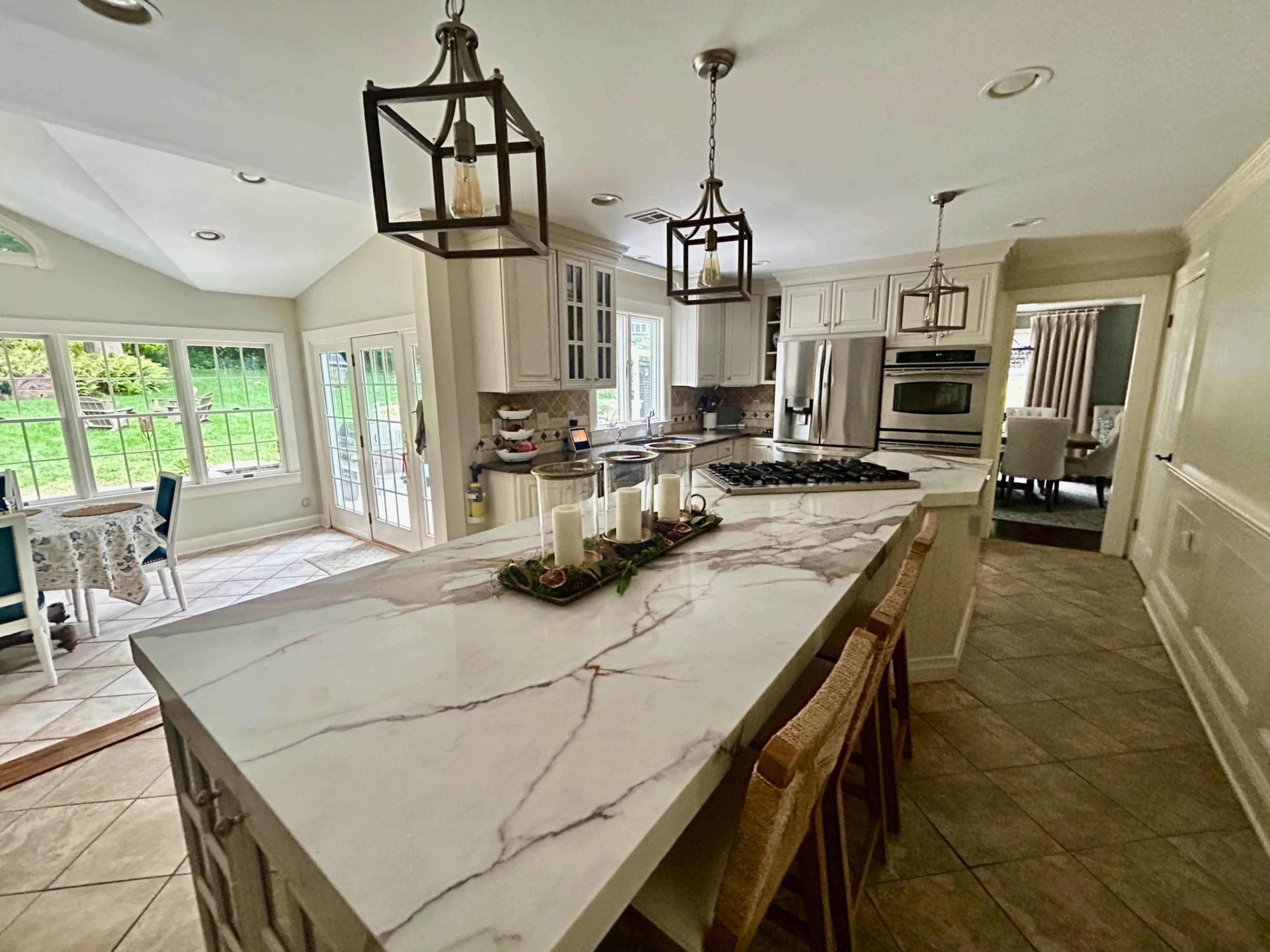 The image shows a modern kitchen with a large marble island, pendant lighting, and an adjacent dining area with a table by the windows.