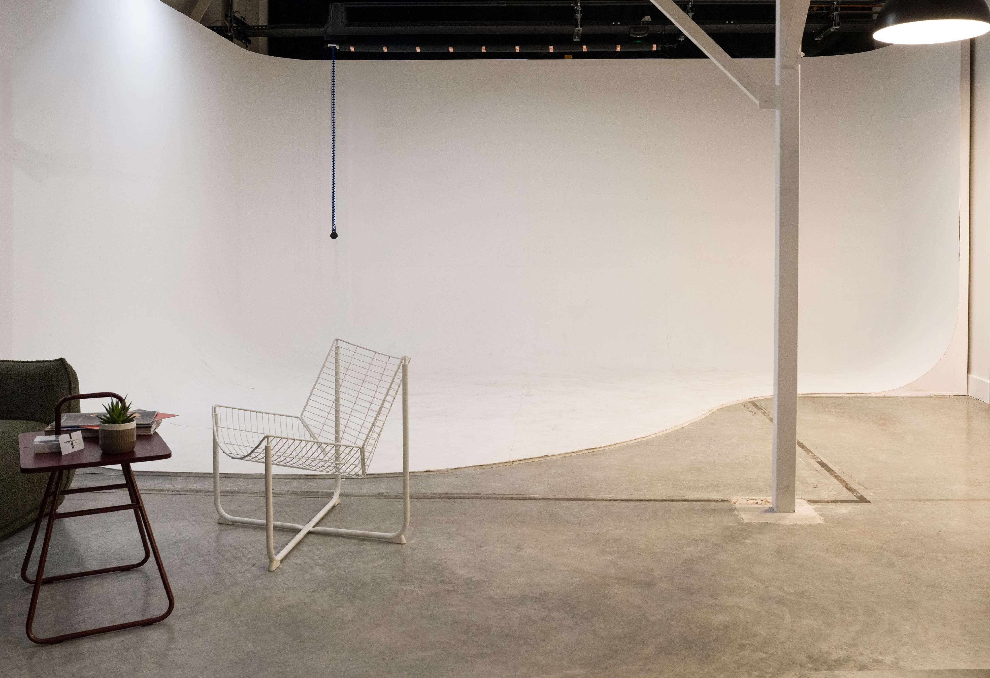 The image shows a minimalist studio space with a smooth white backdrop, a metal chair, and a small table.