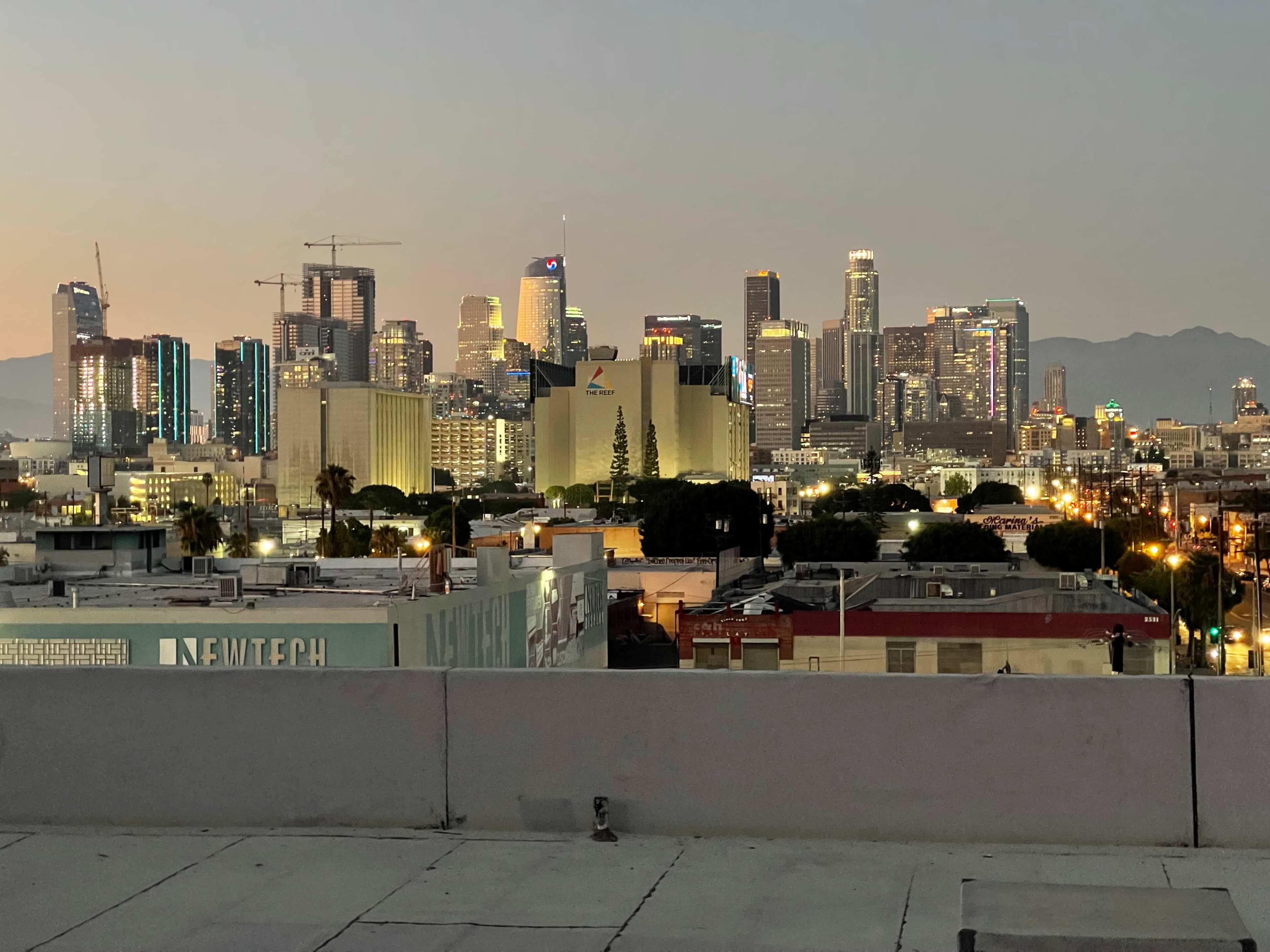 A view of the Los Angeles skyline at dusk, with illuminated buildings and a soft glow in the background.