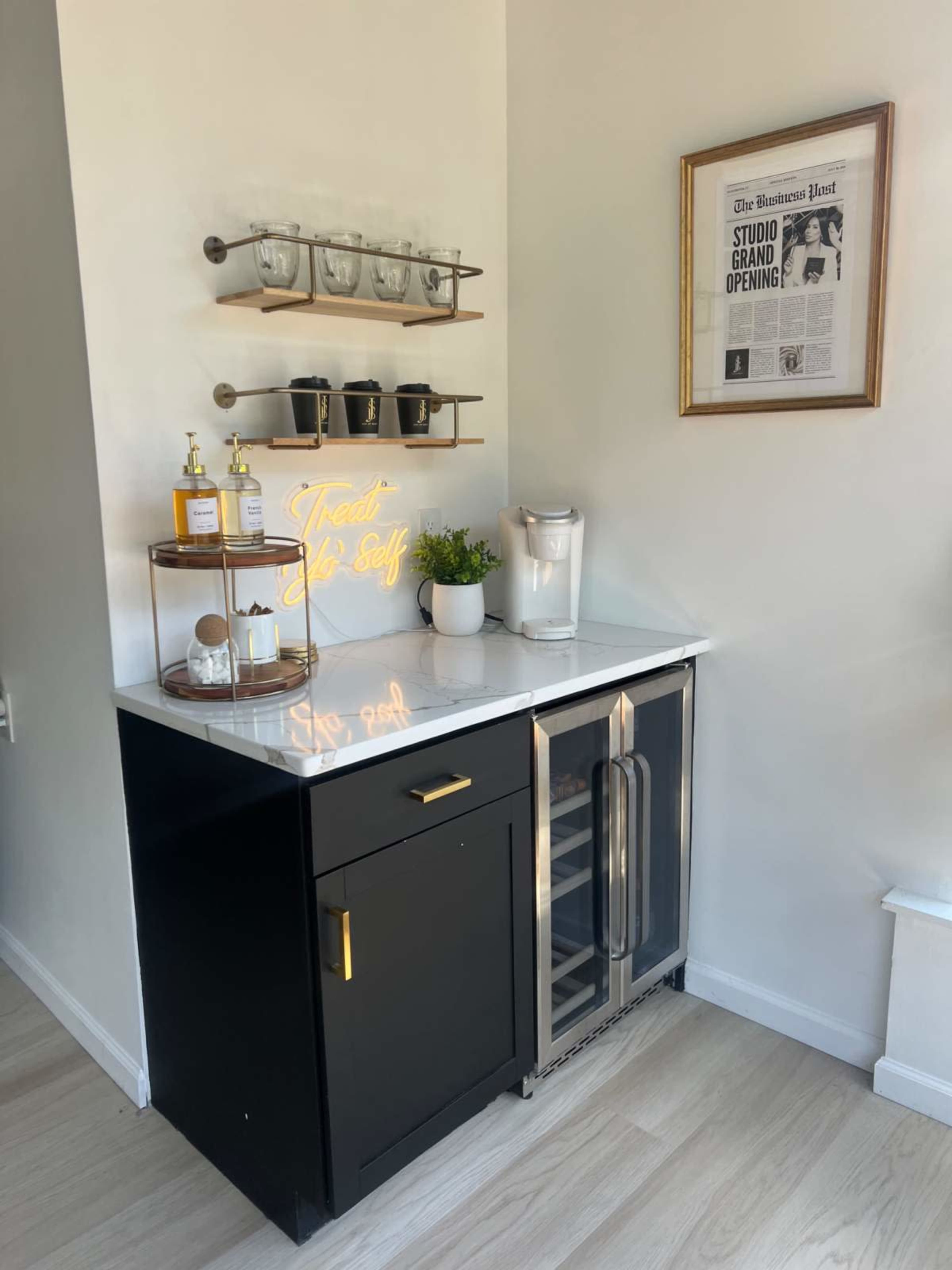 A modern kitchen bar area featuring a black cabinet with a glass door refrigerator, a marble countertop, and a wall-mounted shelf with glassware and decor.