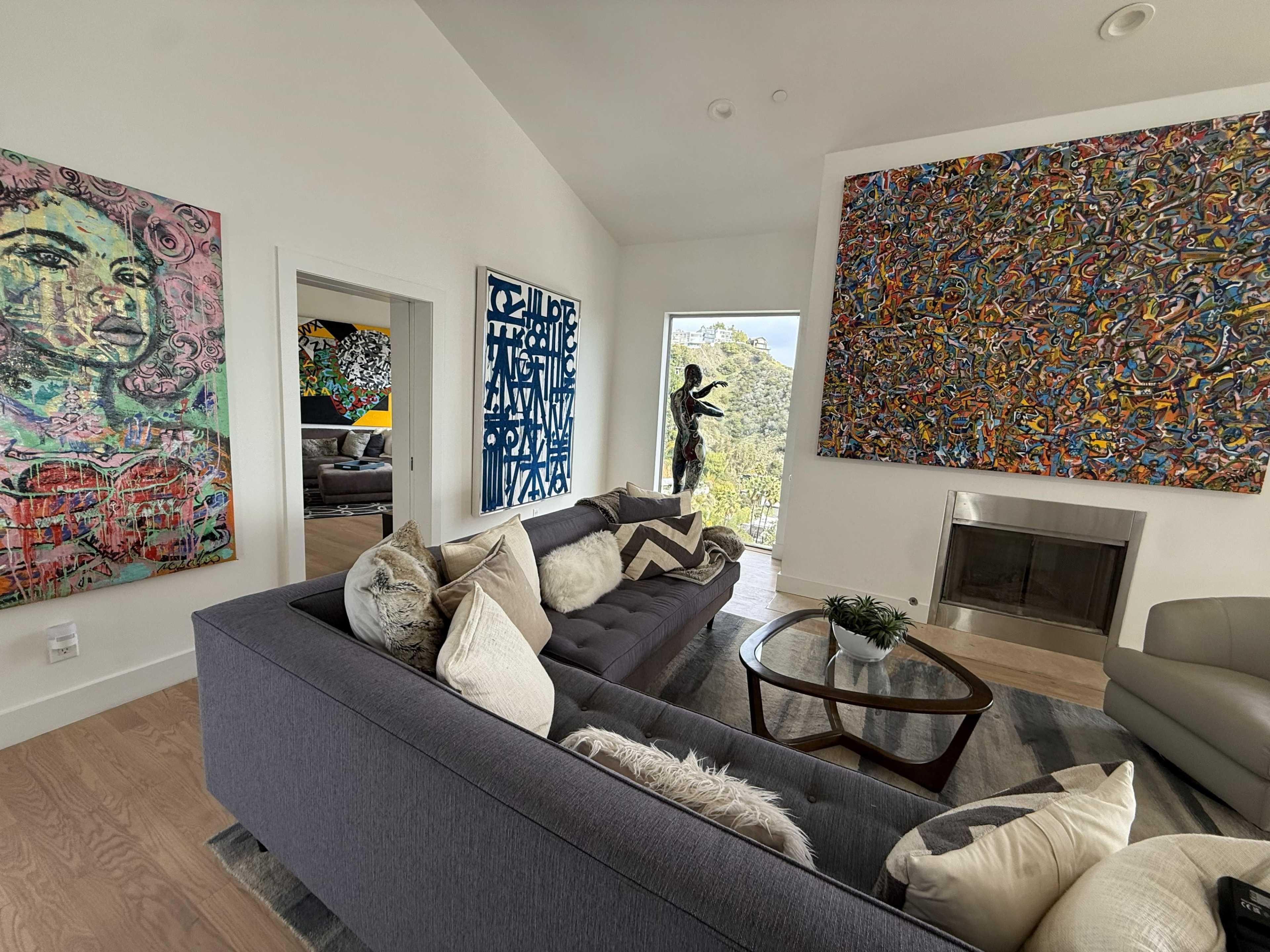 The image shows a modern living room featuring a gray sectional sofa, a round wooden coffee table, and vibrant artwork on the walls, with a view of greenery through a large window.